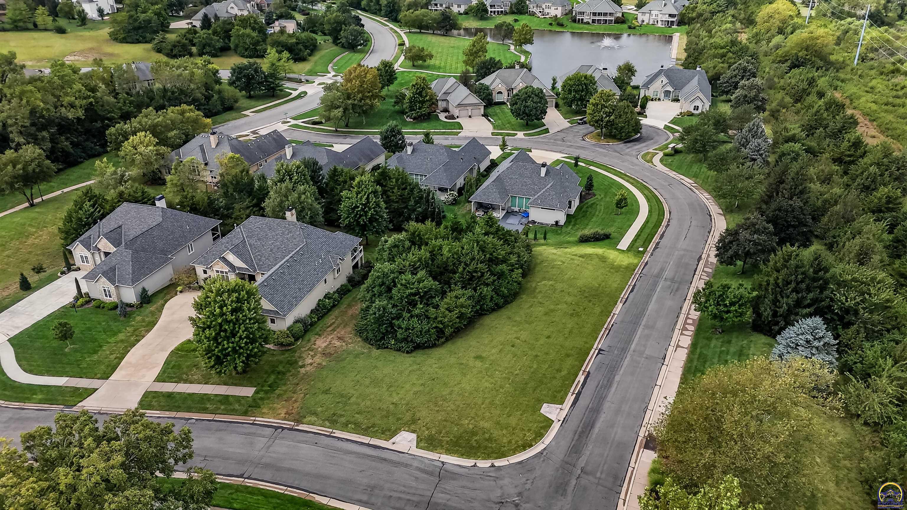 Open grassy corner parcel beside a curving residential street, with homes and a pond nearby.