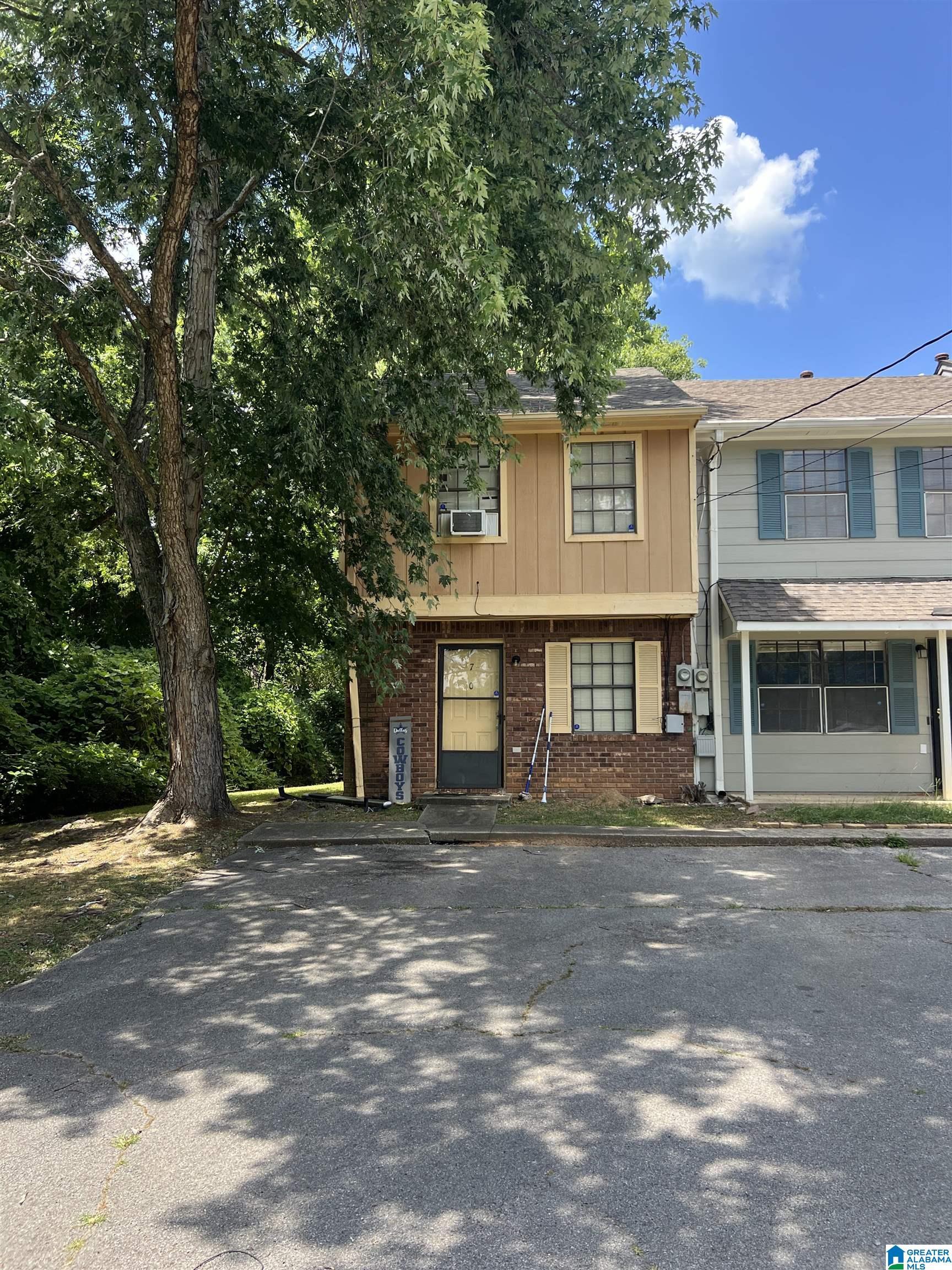 Two-story attached home with brick and tan siding, partly shaded by a large mature tree.