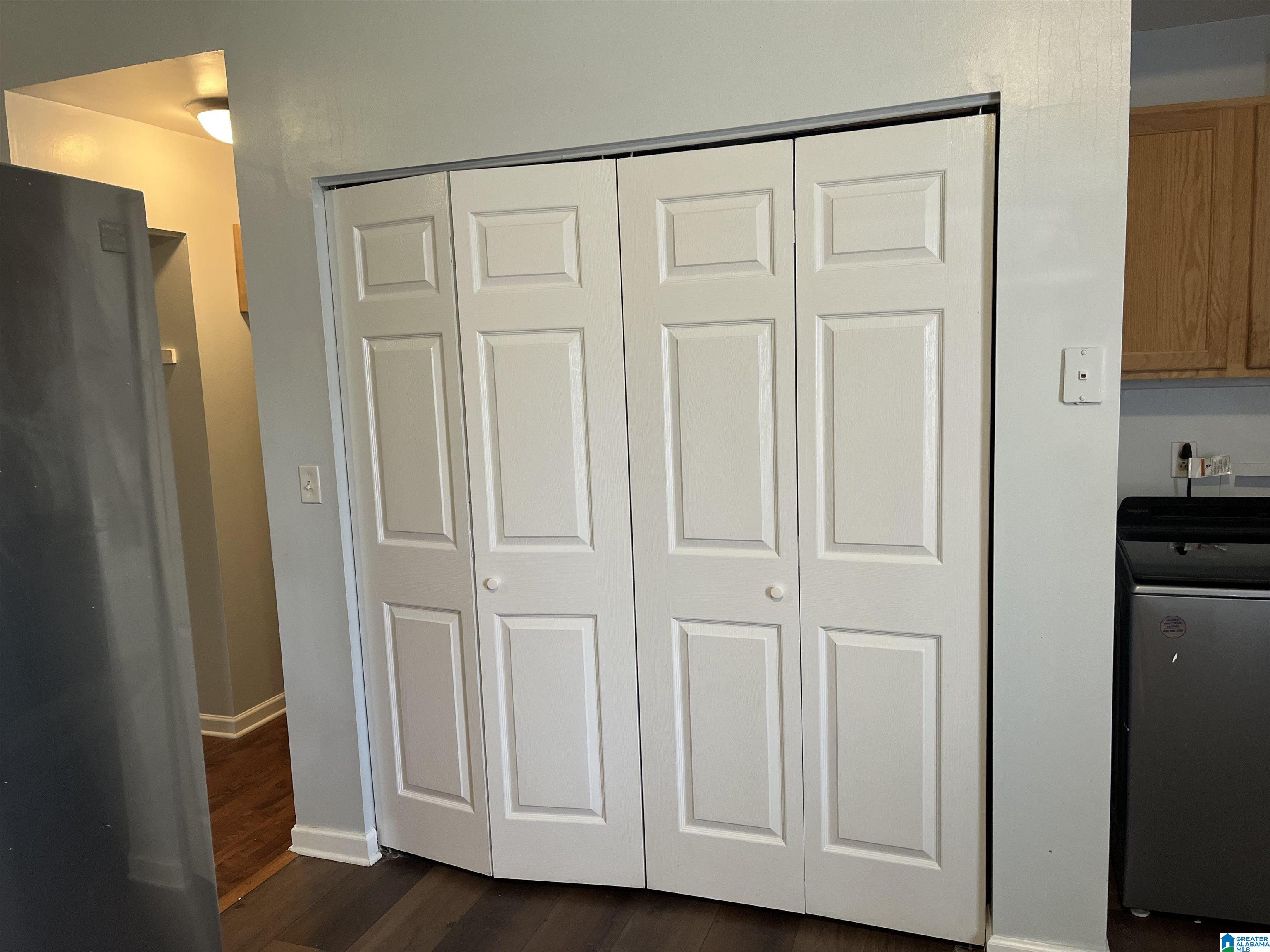 White paneled closet doors between a refrigerator and washer, with wood flooring and kitchen cabinetry nearby.