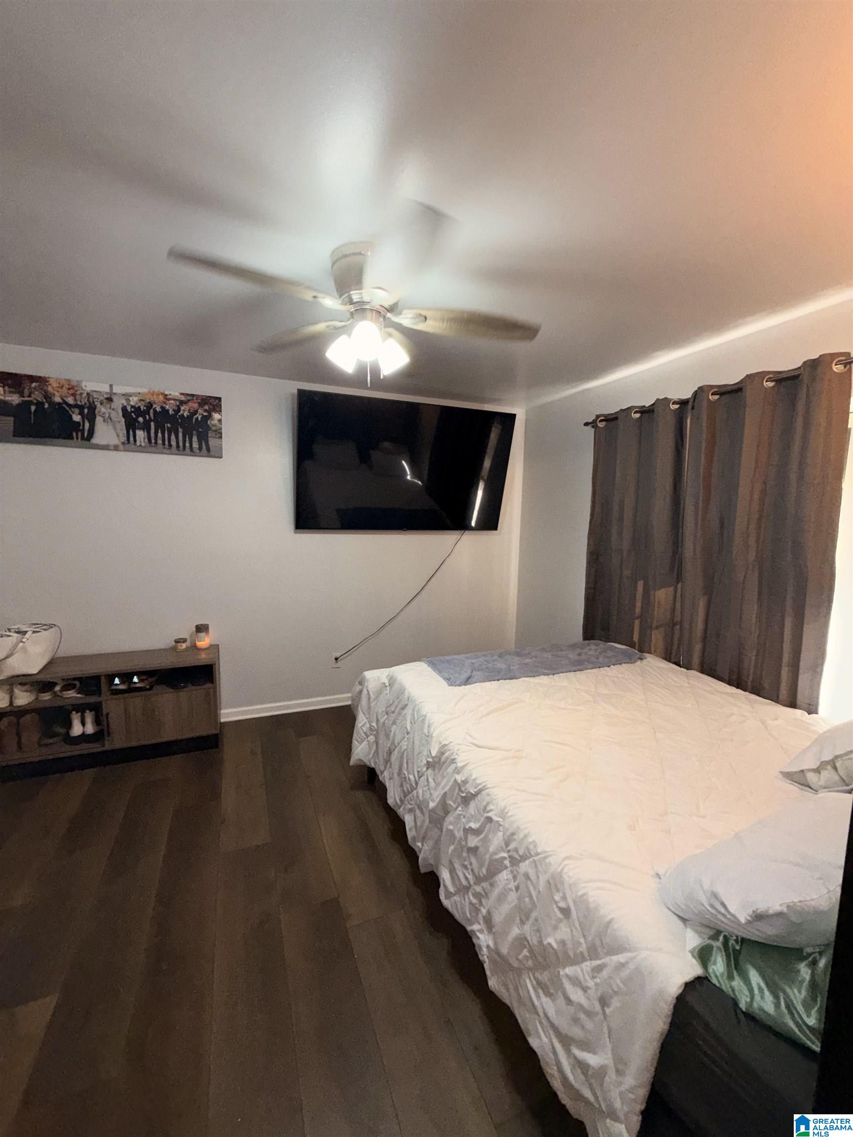 Furnished bedroom with a white bed, dark wood floors, wall-mounted TV, and brown curtains.