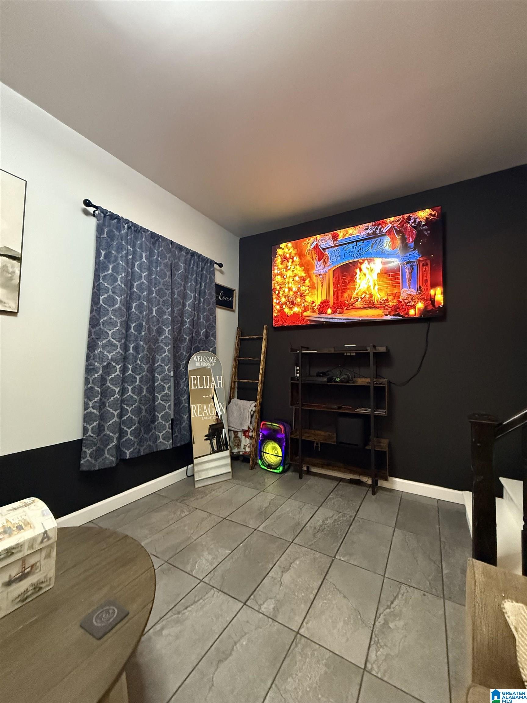 Living room with a wall-mounted television, dark accent wall, patterned curtains, and gray tile flooring.