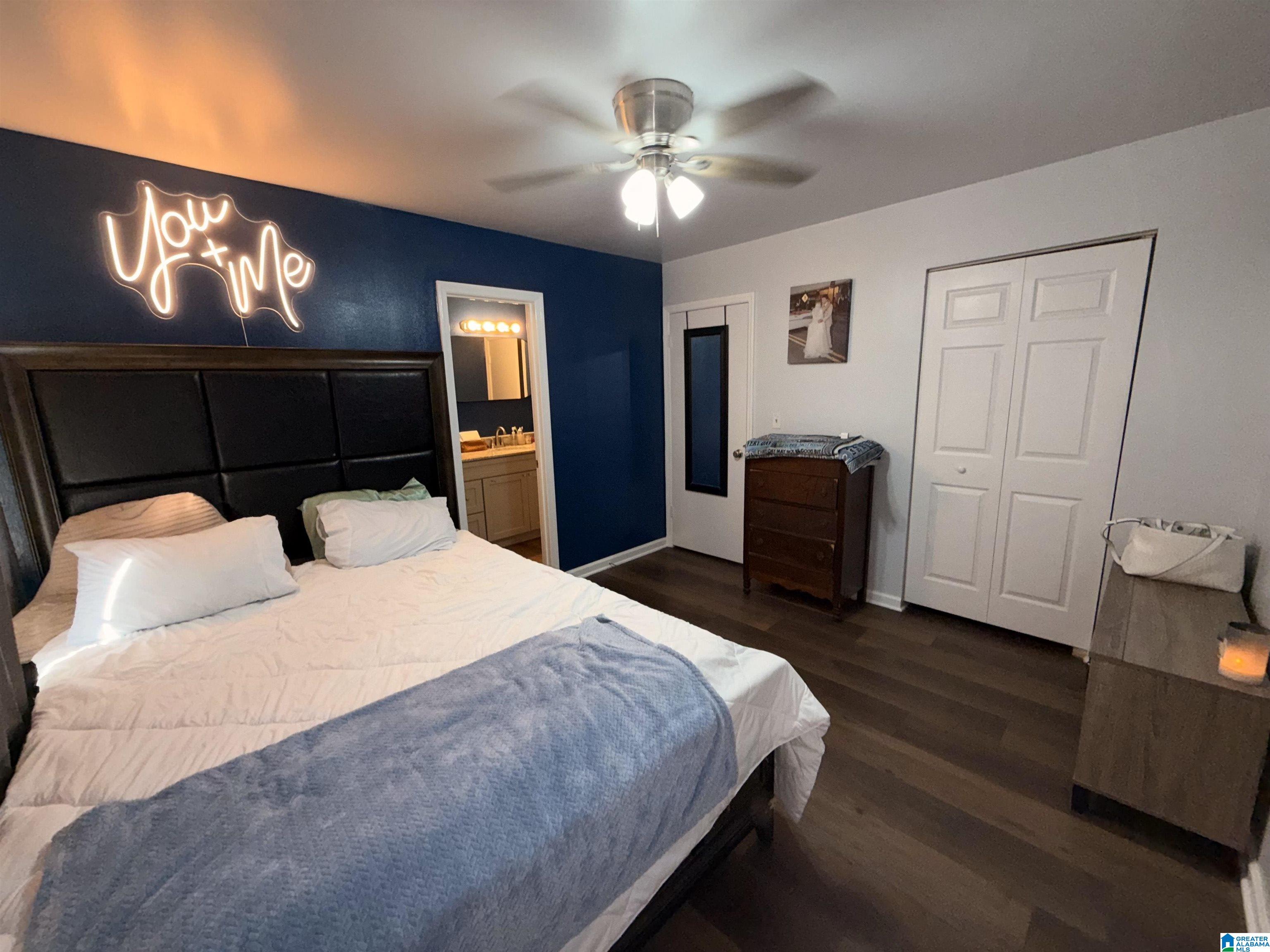 Bedroom with a large bed, navy accent wall, glowing “You + Me” sign, and doorway to a bathroom vanity.
