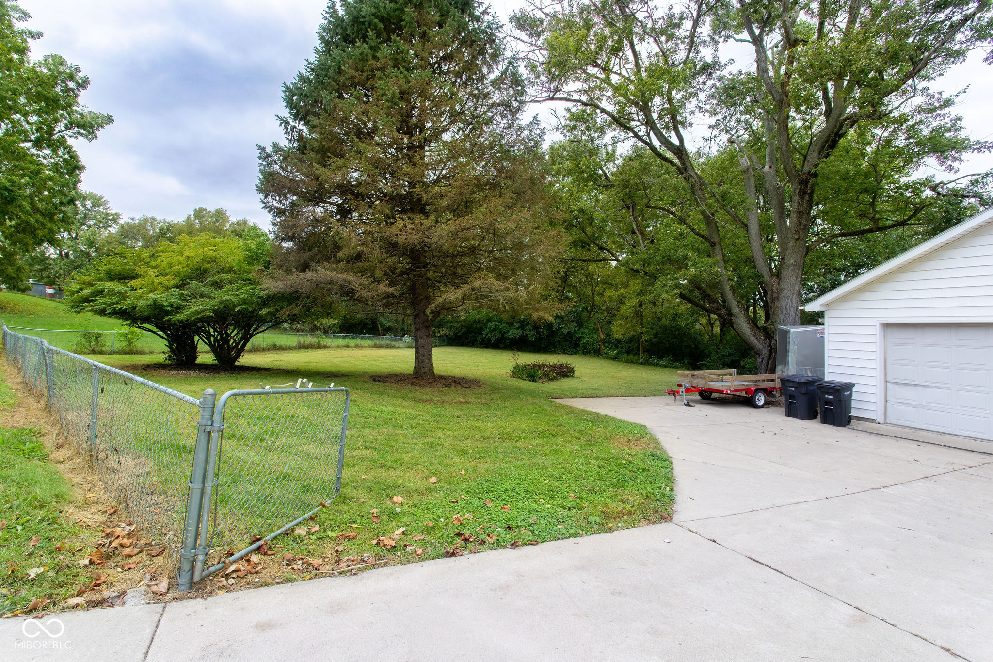 Grassy backyard with mature trees, chain-link fence, and a driveway beside a white garage.