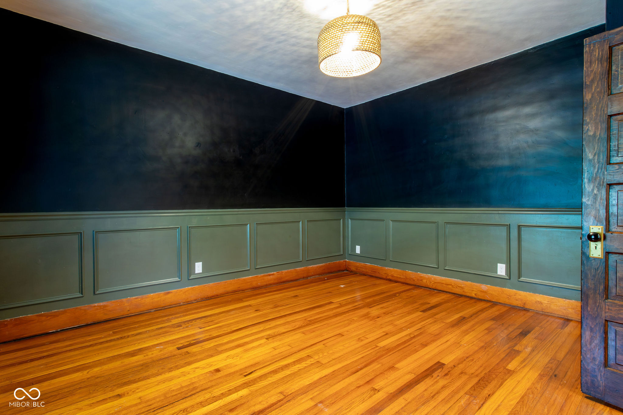 Empty room with hardwood floors, dark blue walls, paneled wainscoting, and a woven pendant light.