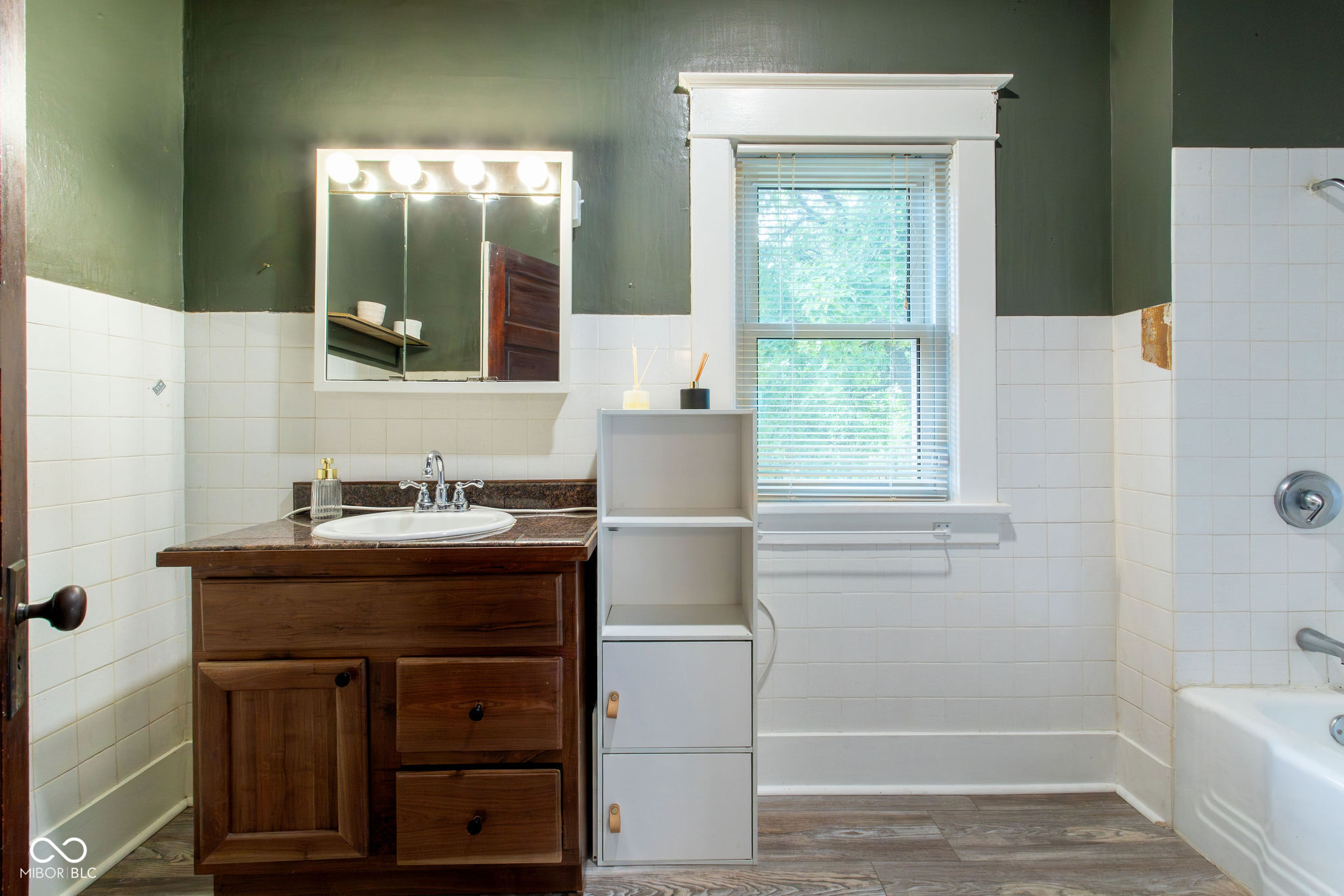 Bathroom with a dark wood vanity, lit mirrored cabinet, window, and white-tiled bathtub surround.