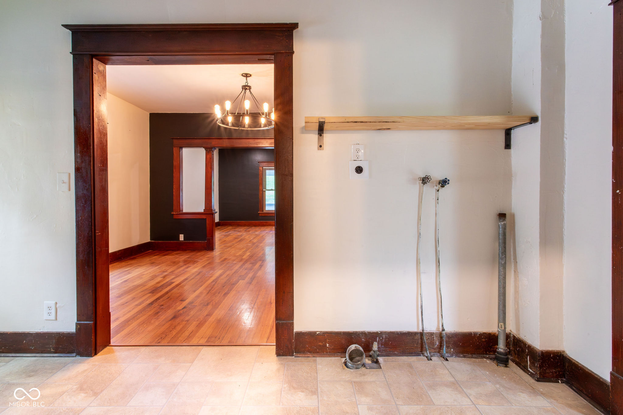 Vacant utility area with exposed plumbing and a wooden shelf beside a dark-trimmed doorway.