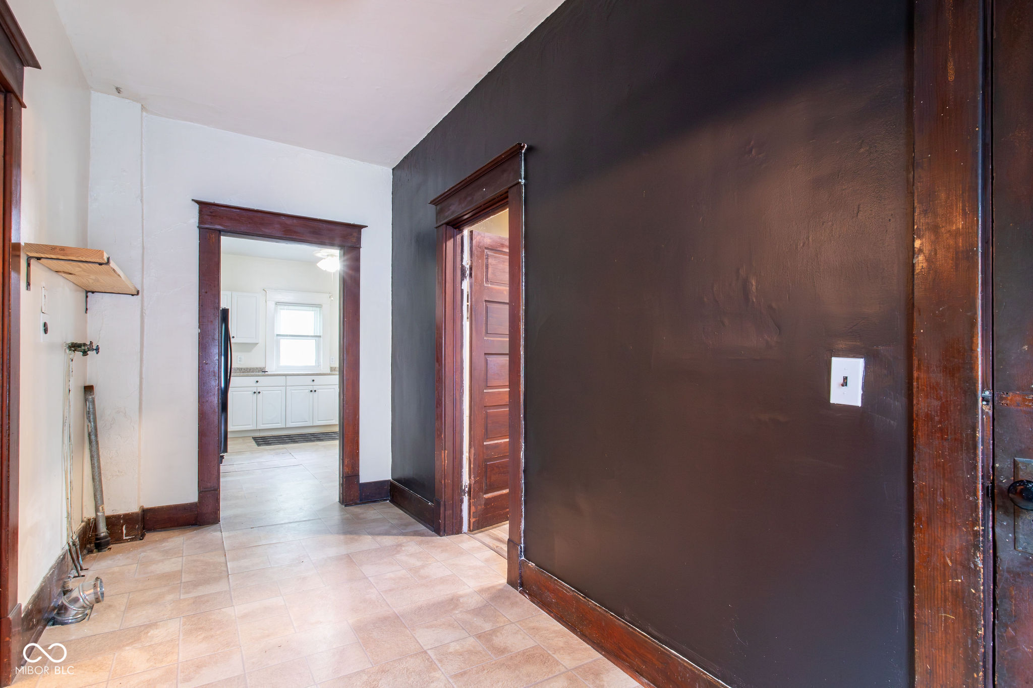 Hallway with tiled floors and dark wood trim, looking through to a bright white kitchen.