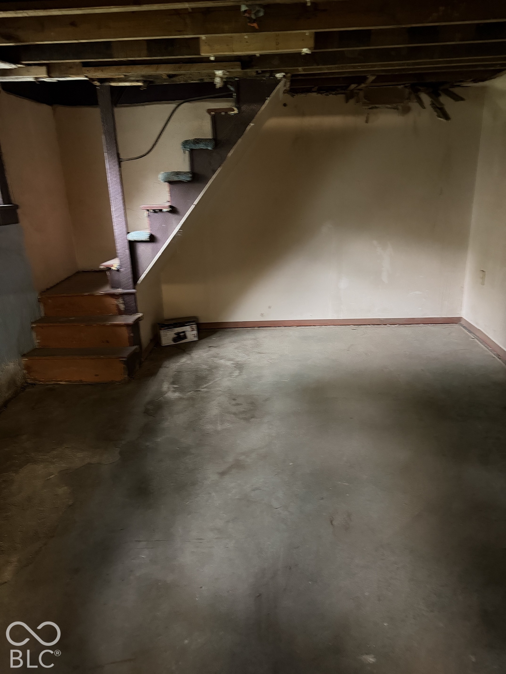 Empty basement with a concrete floor, exposed ceiling joists, and wooden stairs.