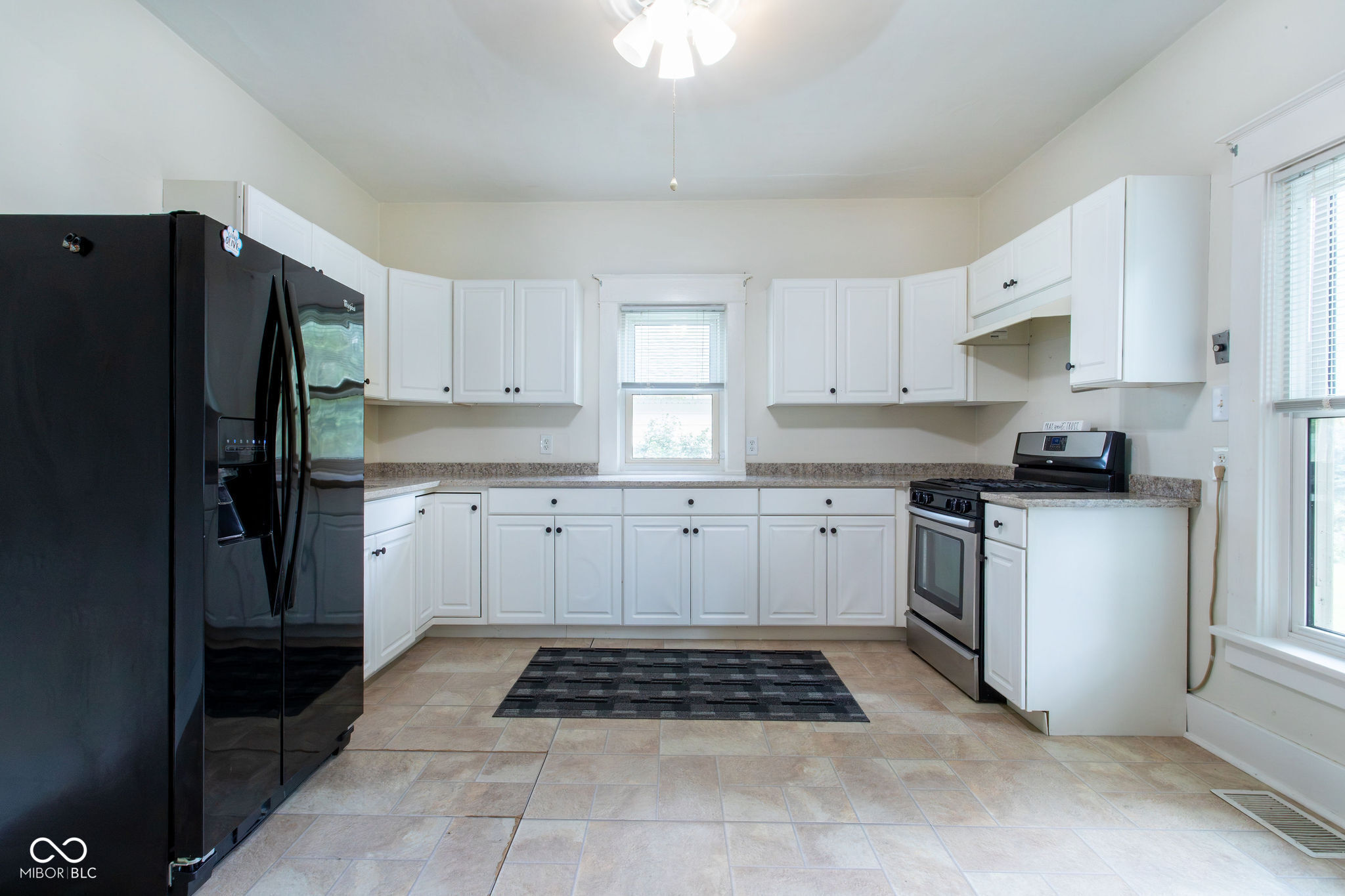 Kitchen with white cabinets, black appliances, beige tile floors, and windows above the counter.