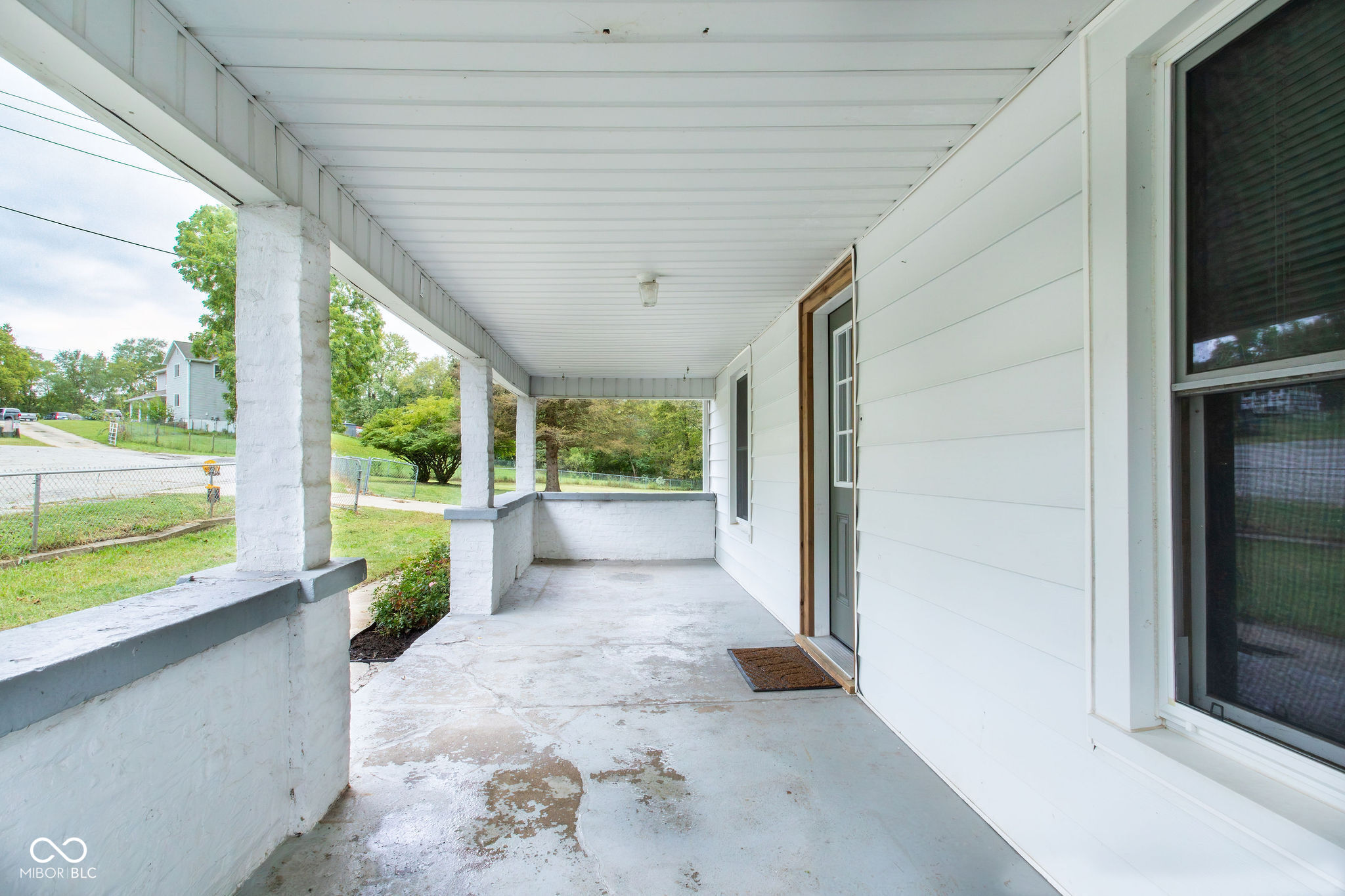 Covered concrete front porch with white columns, siding, and a view of the fenced lawn.