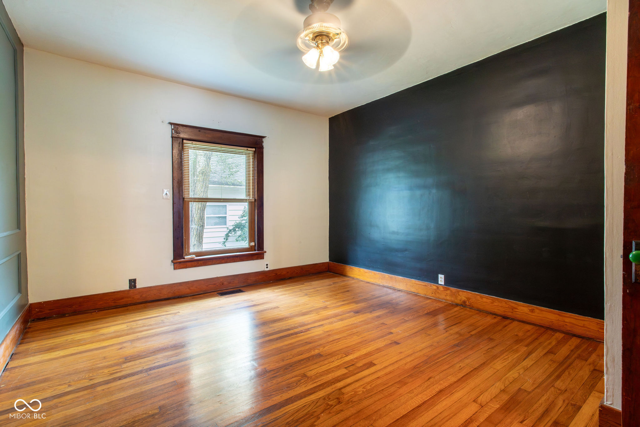 Empty room with polished hardwood floors, a dark accent wall, and a wood-trimmed window.