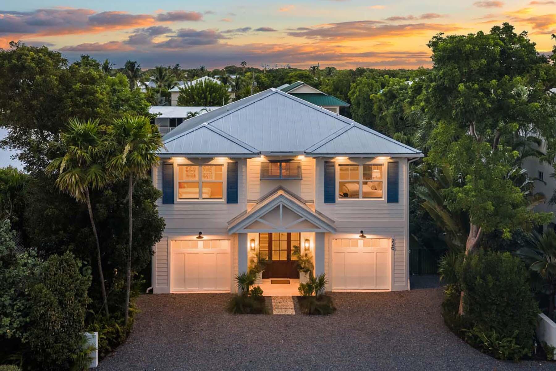White two-story house with blue shutters and a gravel driveway, warmly lit beneath a sunset sky.