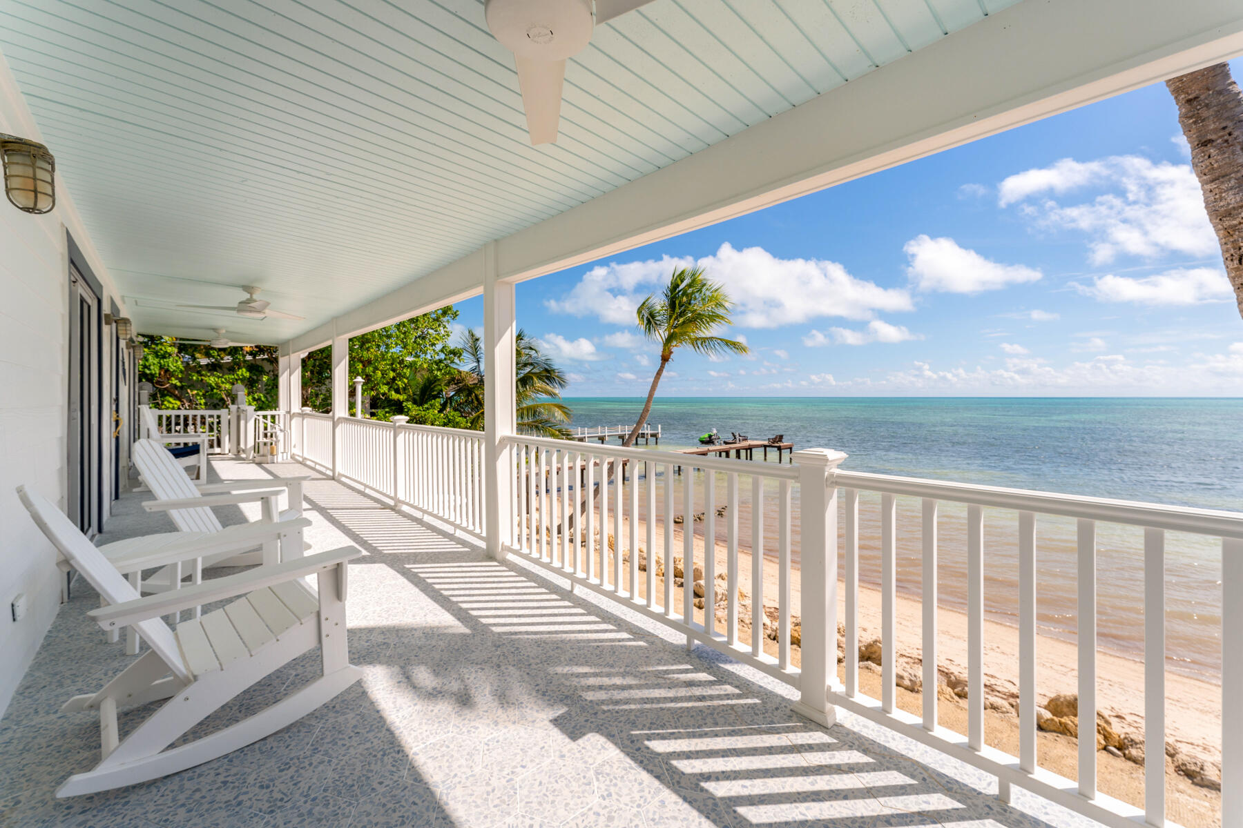 Covered waterfront porch with white Adirondack chairs overlooking a sandy beach and turquoise ocean.