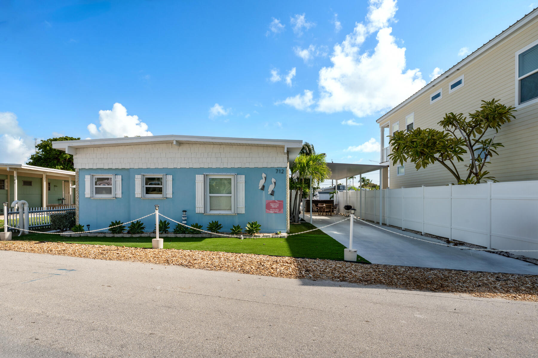 Blue cottage with white trim and shutters, numbered 712, beside a white fence and driveway.