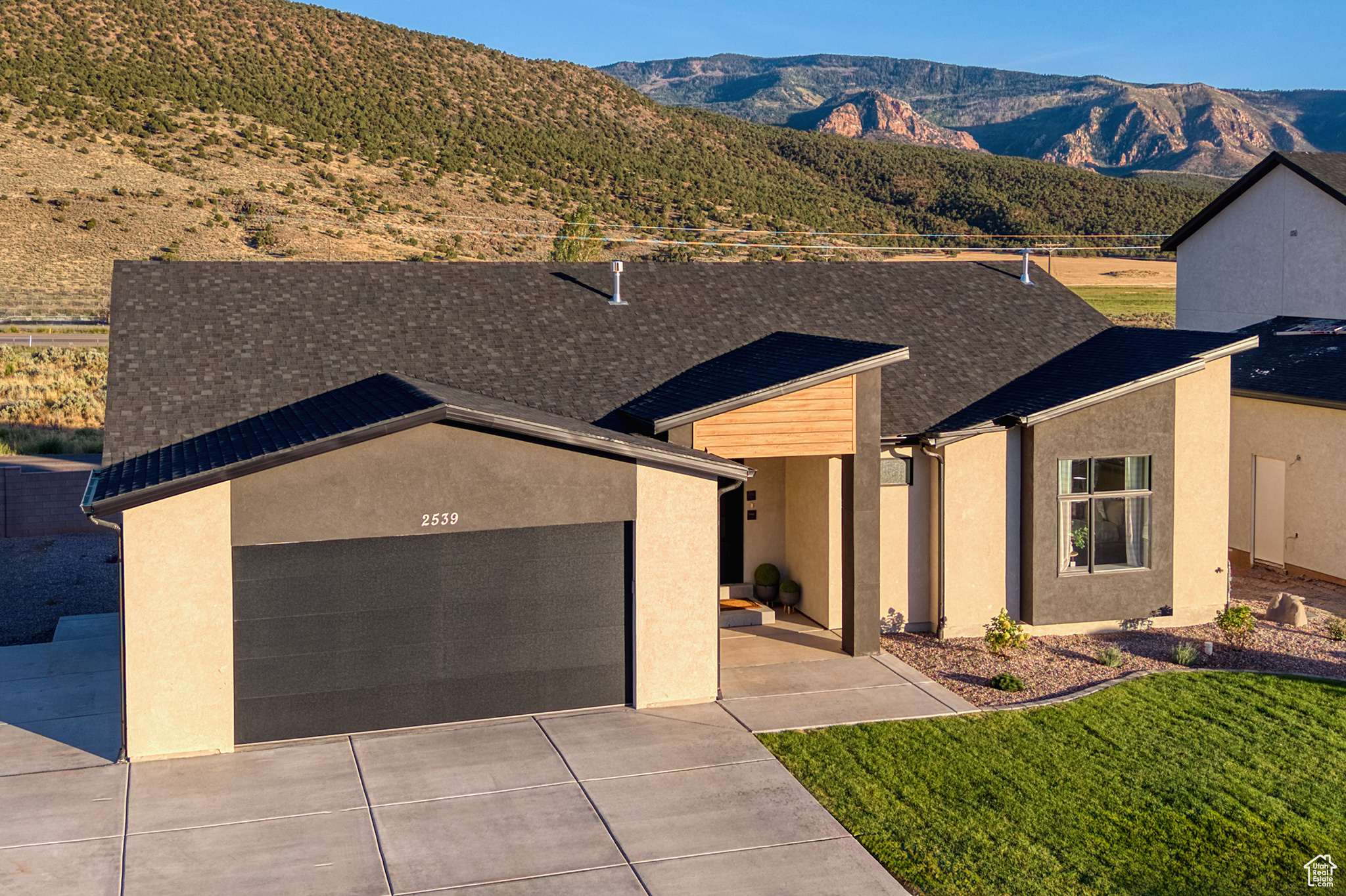 Modern home with dark double garage door and covered entry, backed by mountains.