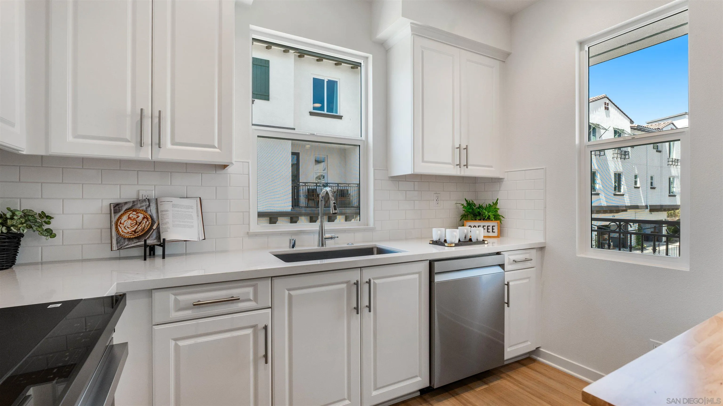 Bright white kitchen with quartz counters, subway tile backsplash, stainless dishwasher, and windows overlooking neighboring homes.