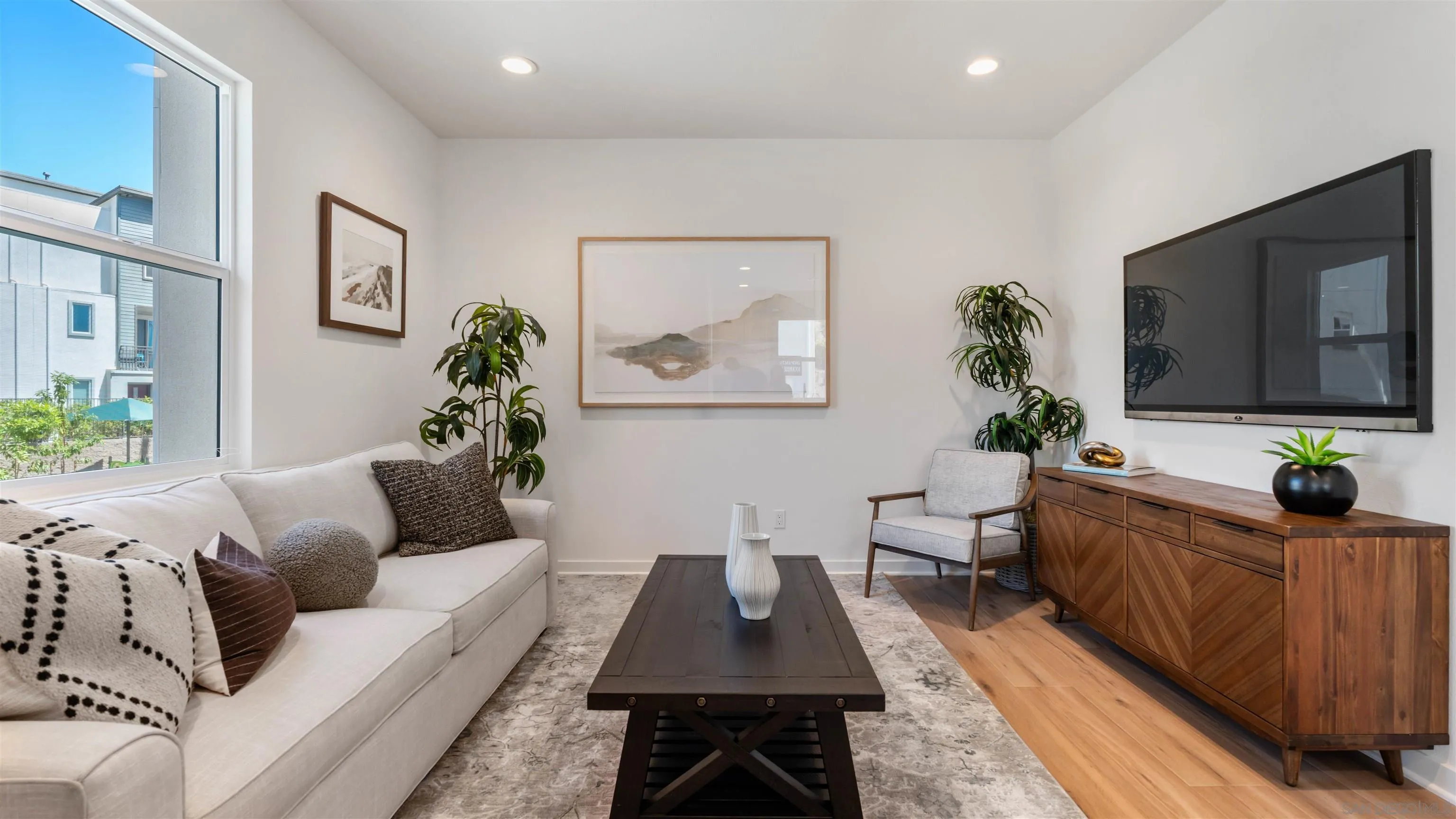 Staged living room with a white sofa, large window, and wood media console beneath a wall-mounted television.