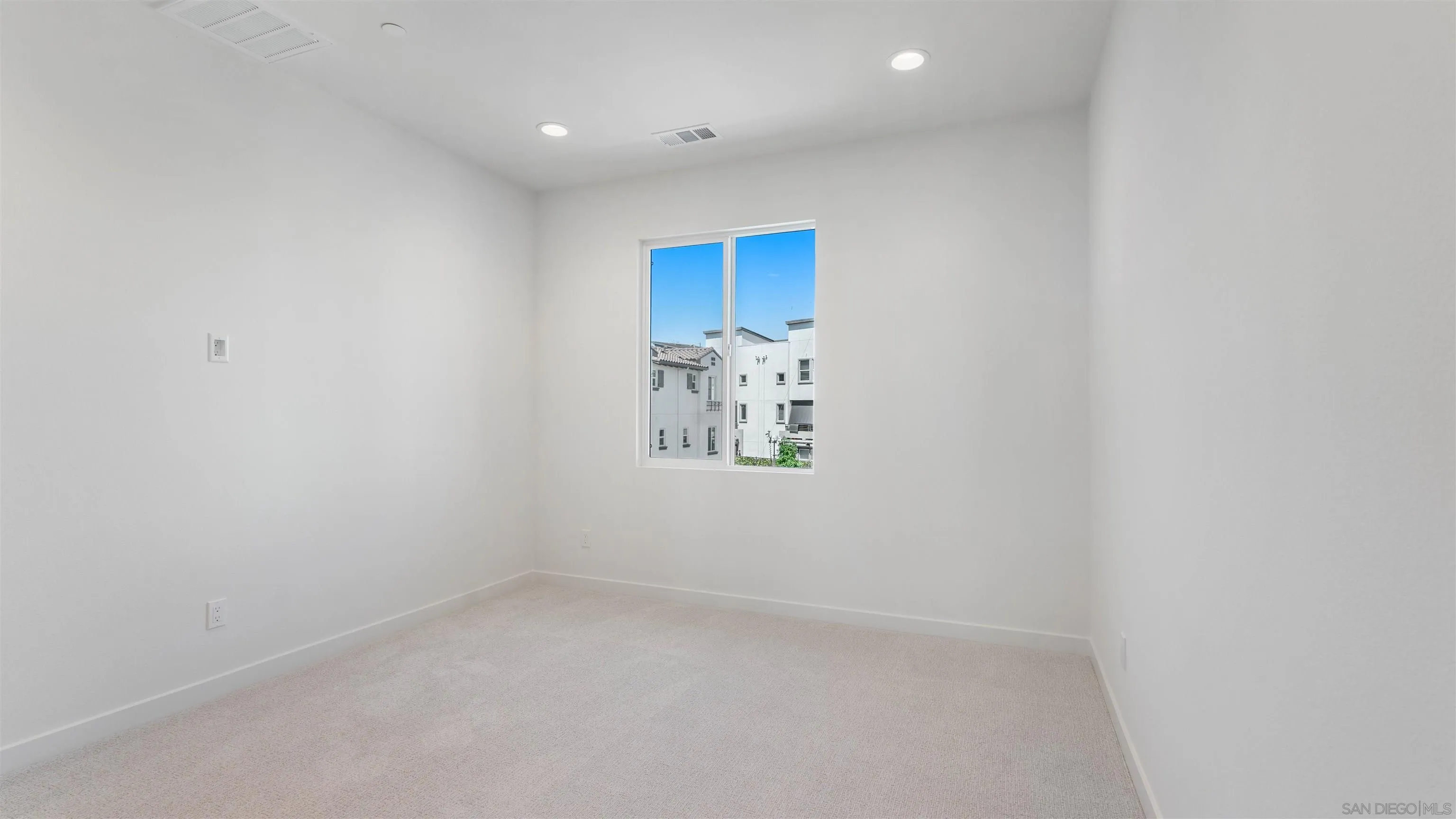 Empty carpeted bedroom with recessed lights and a window overlooking neighboring homes.