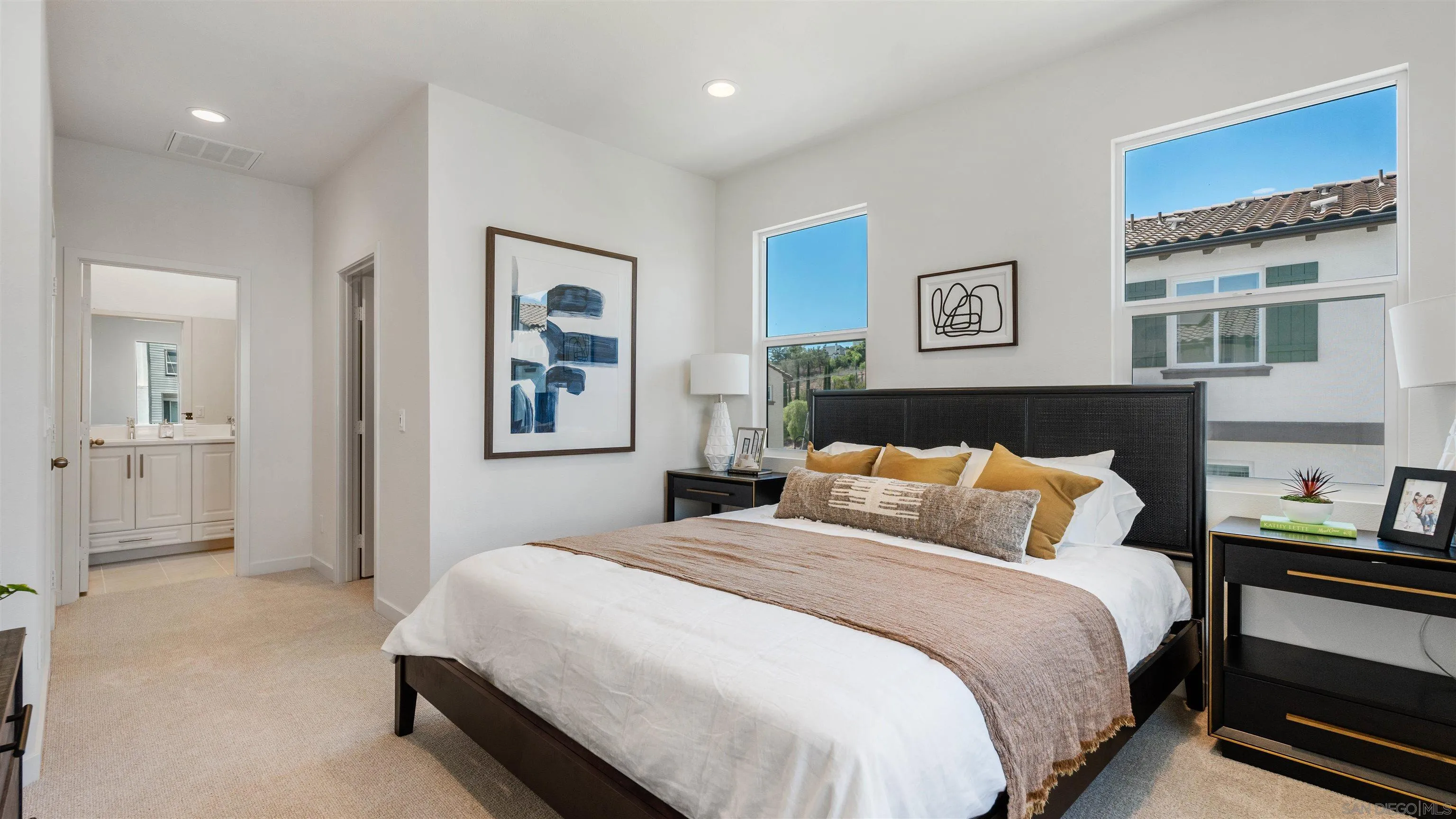 Staged bedroom with a dark bed, ochre pillows, tall windows, and an open doorway to the ensuite bathroom.