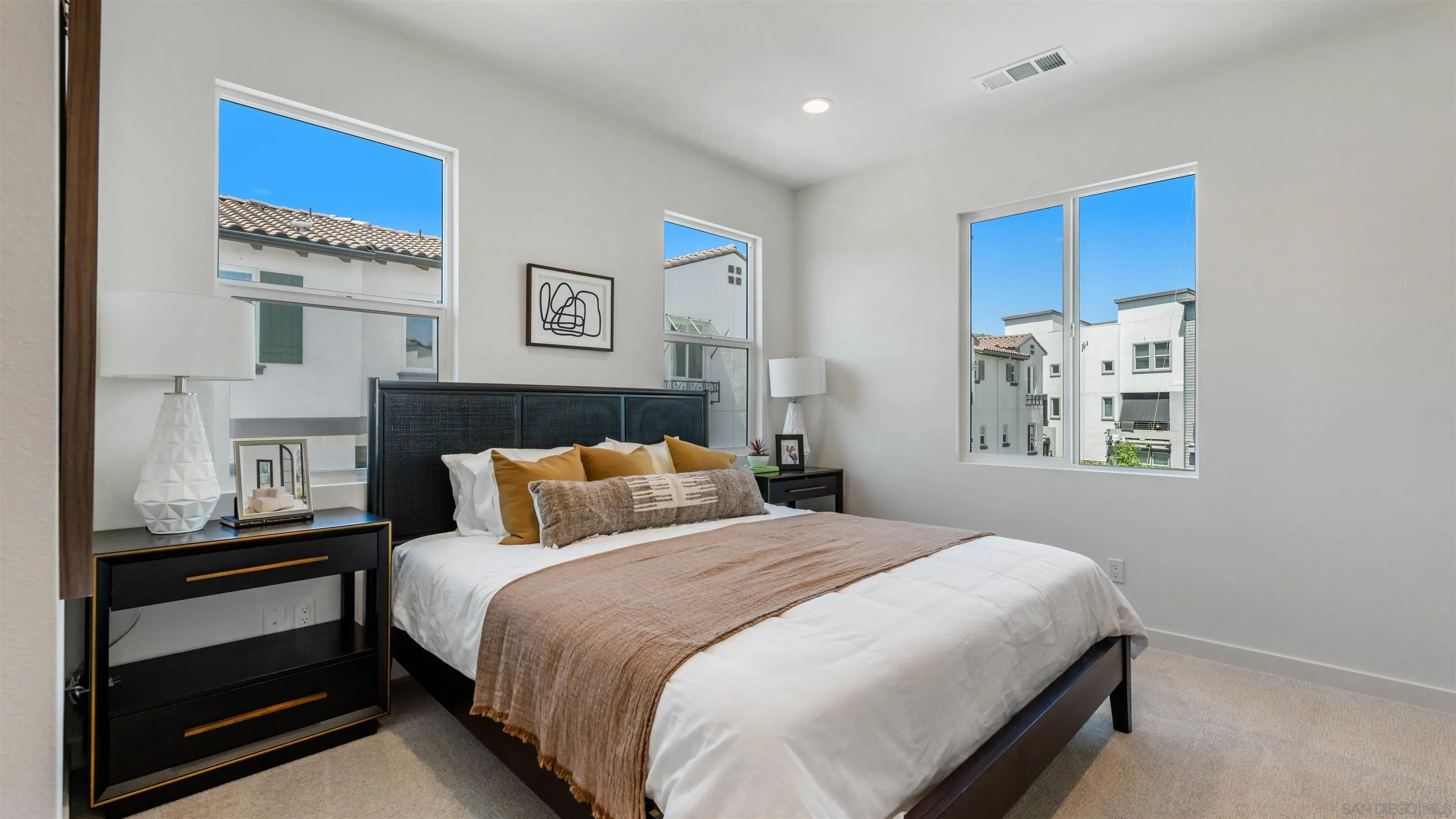 Staged bedroom with a large white bed, dark bedside furniture, and tall windows overlooking neighboring homes.