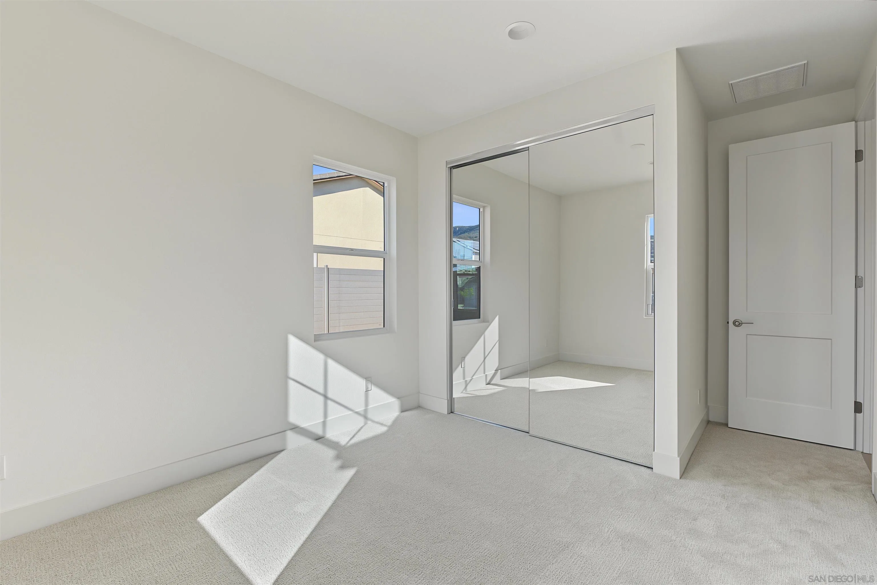 Empty carpeted bedroom with mirrored closet doors and sunlight streaming through the window.