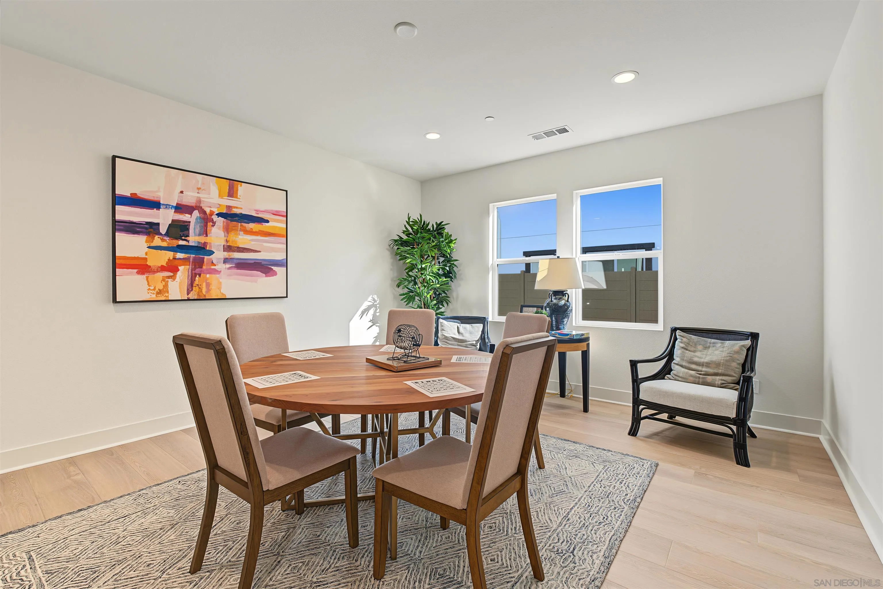 Dining room with a round wooden table, upholstered chairs, colorful abstract art, and two windows.