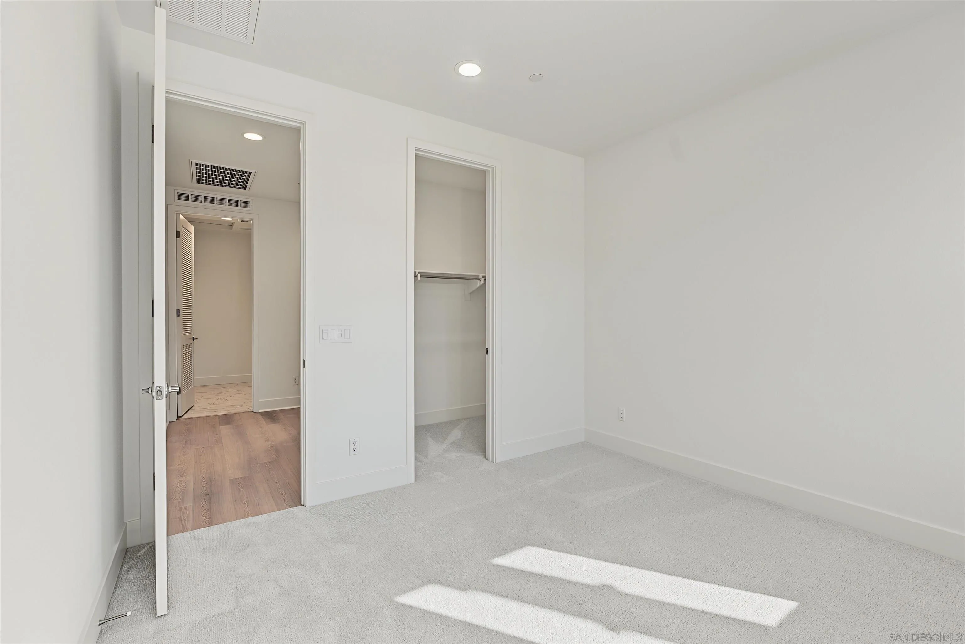 Empty carpeted bedroom with an open closet and doorway to a wood-floored hall.