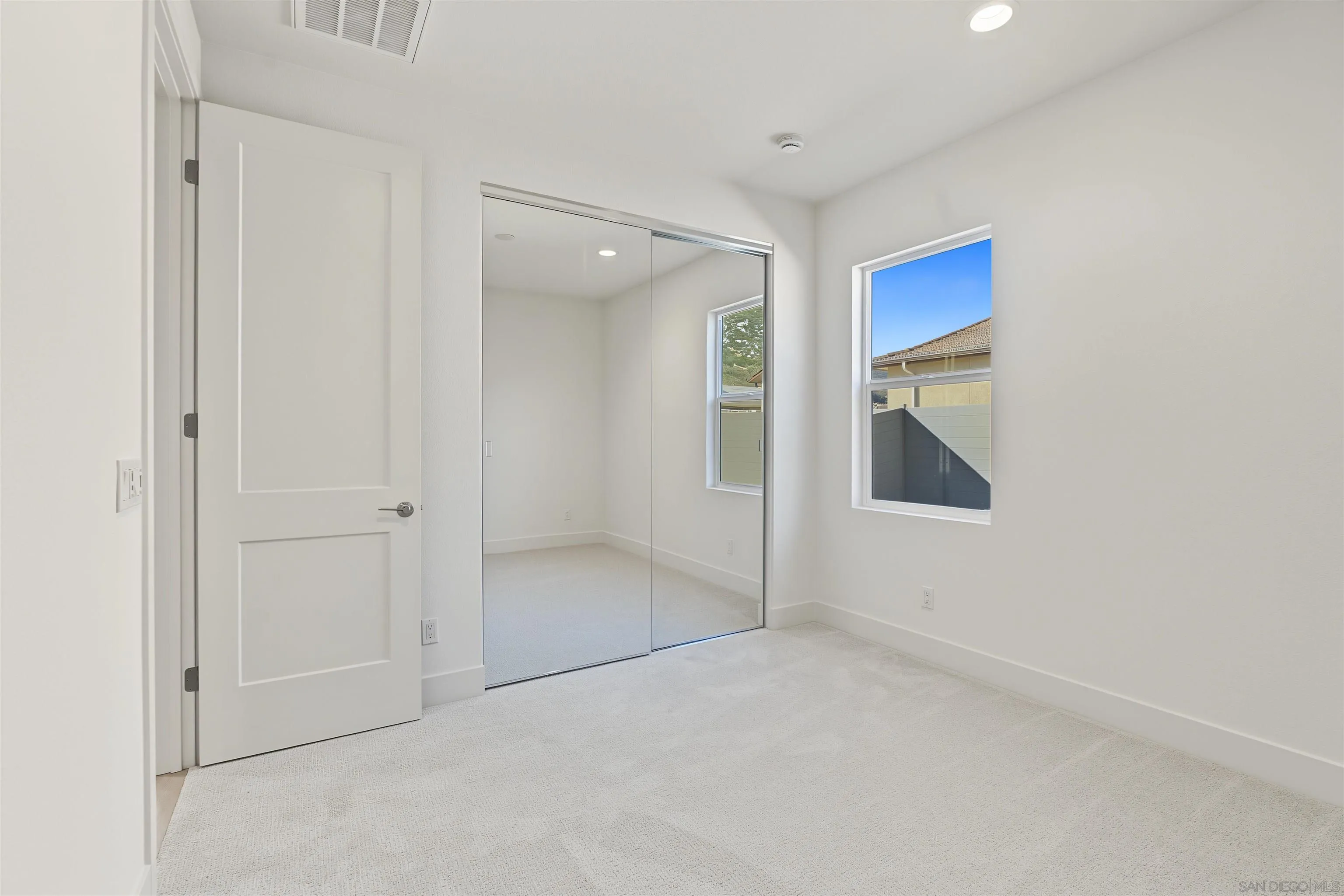 Empty bedroom with mirrored closet doors, light carpet, and a window overlooking a neighboring home.