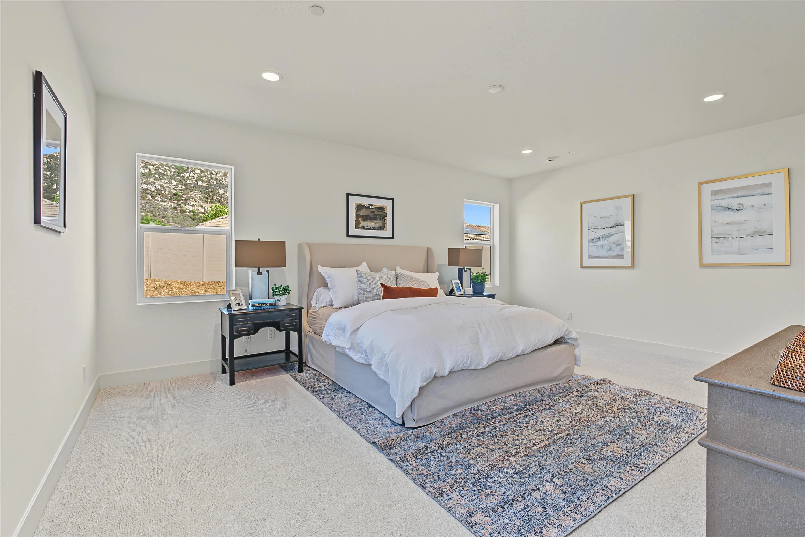 Staged bedroom with a white bed, patterned blue rug, and windows overlooking a hillside.