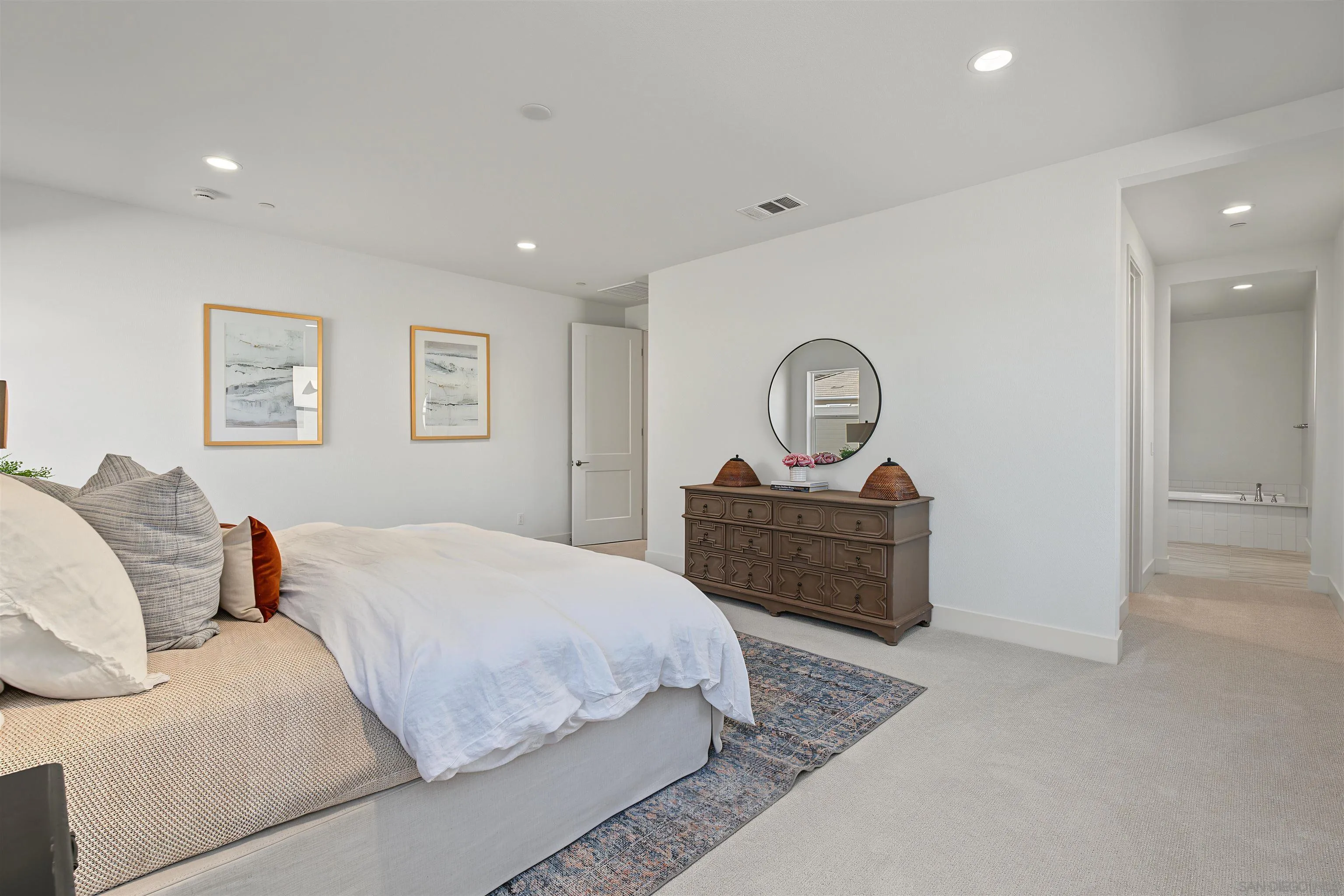 Staged bedroom with a large bed, patterned rug, and dark wood dresser beneath a round mirror.