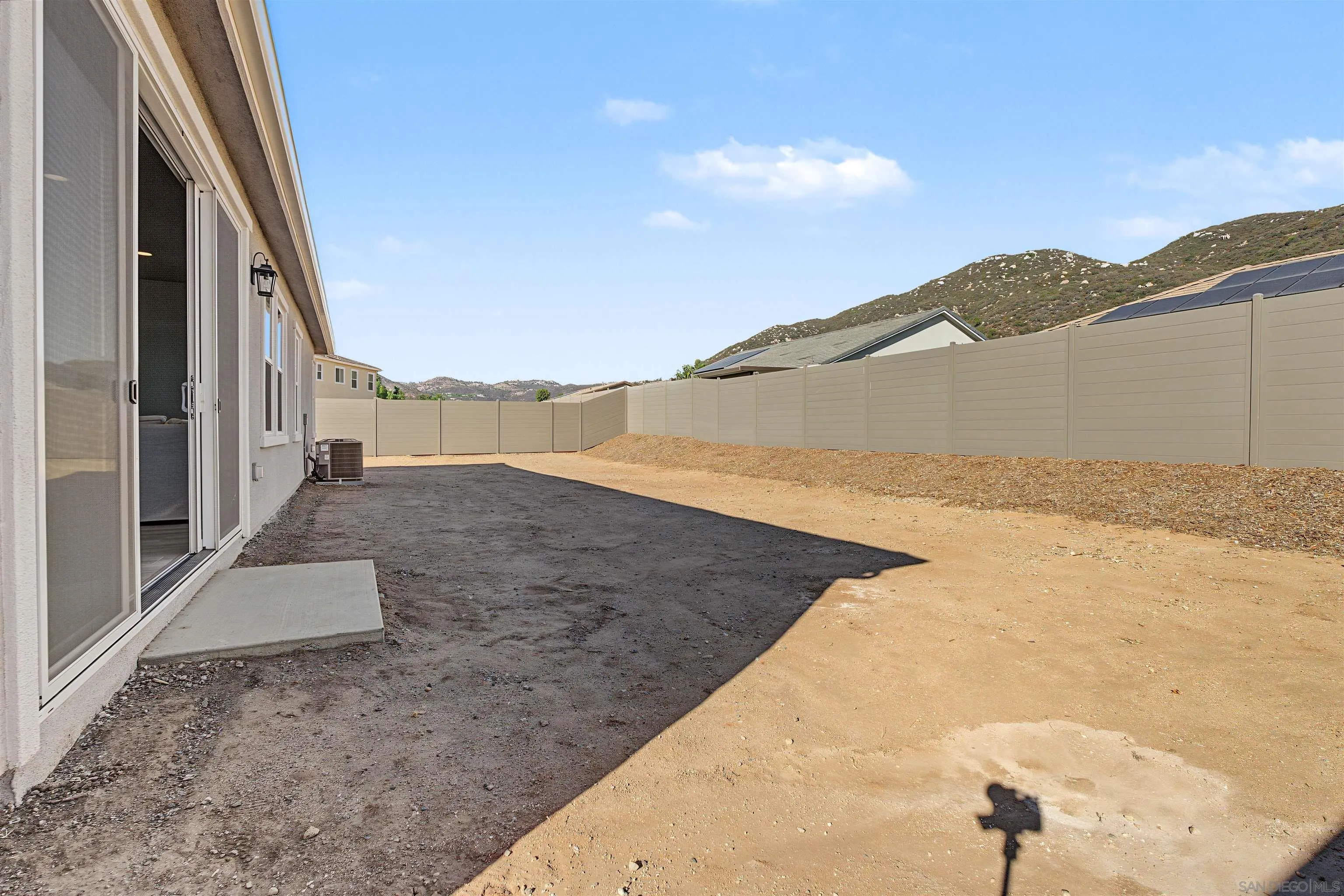 Bare dirt backyard beside the house, enclosed by a tall privacy fence with low hills beyond.