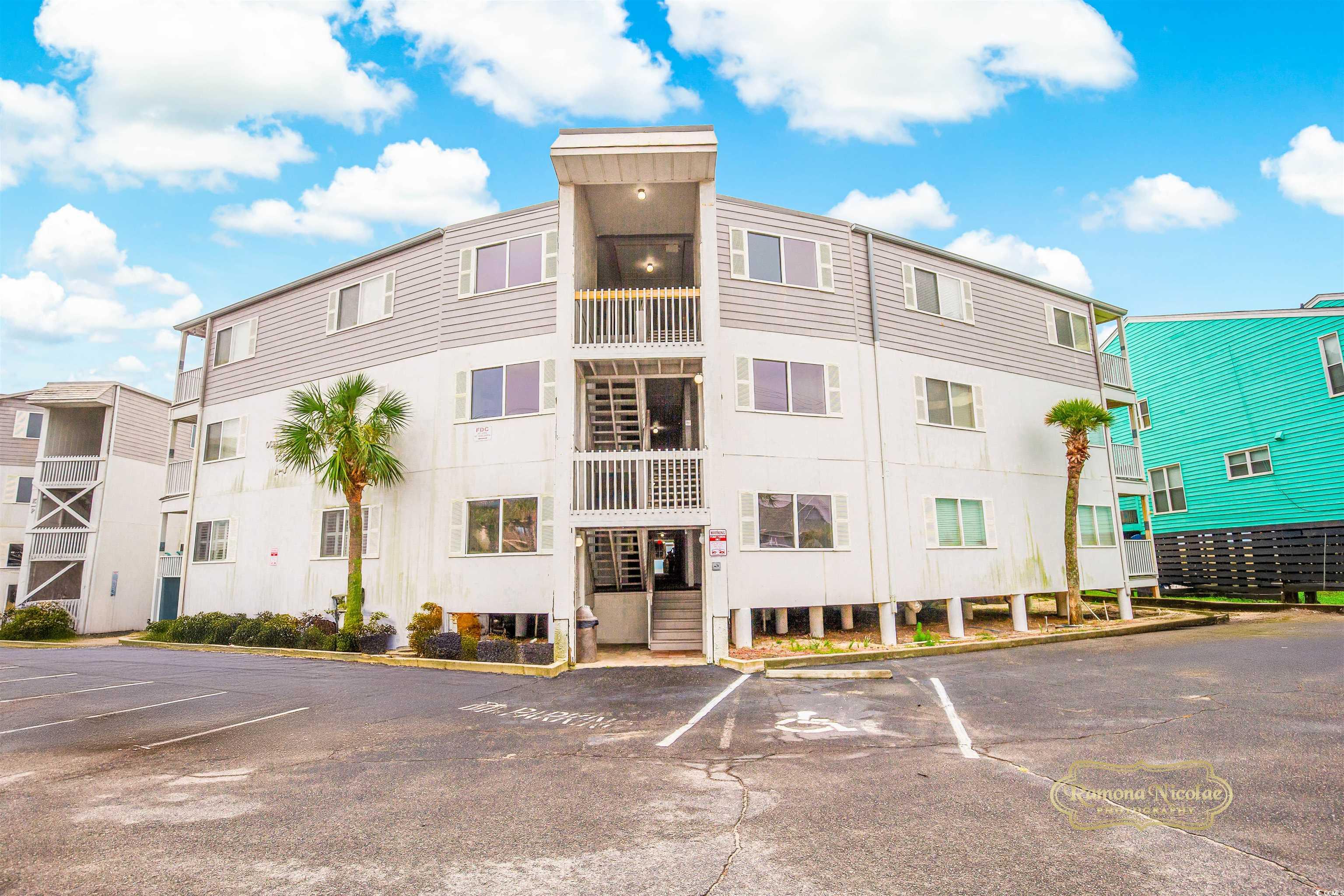 Three-story condominium building with central stairway, balconies, palm trees, and an accessible parking space.