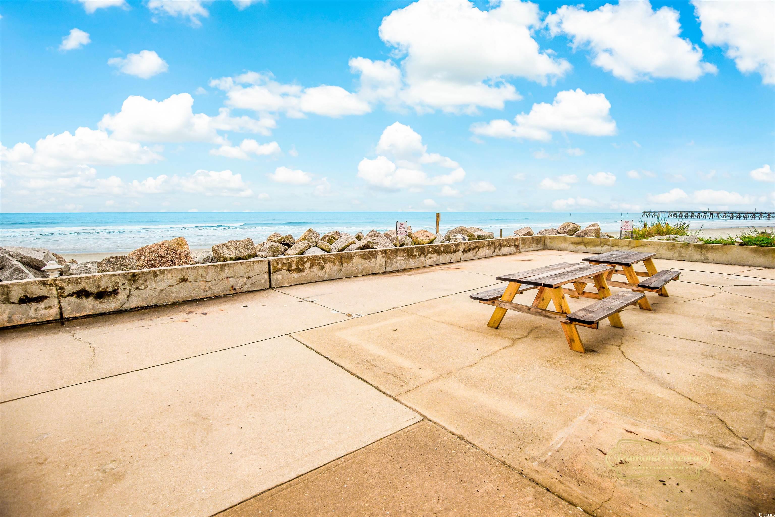 Oceanfront patio with wooden picnic tables overlooking the beach, rocky seawall, and distant pier.