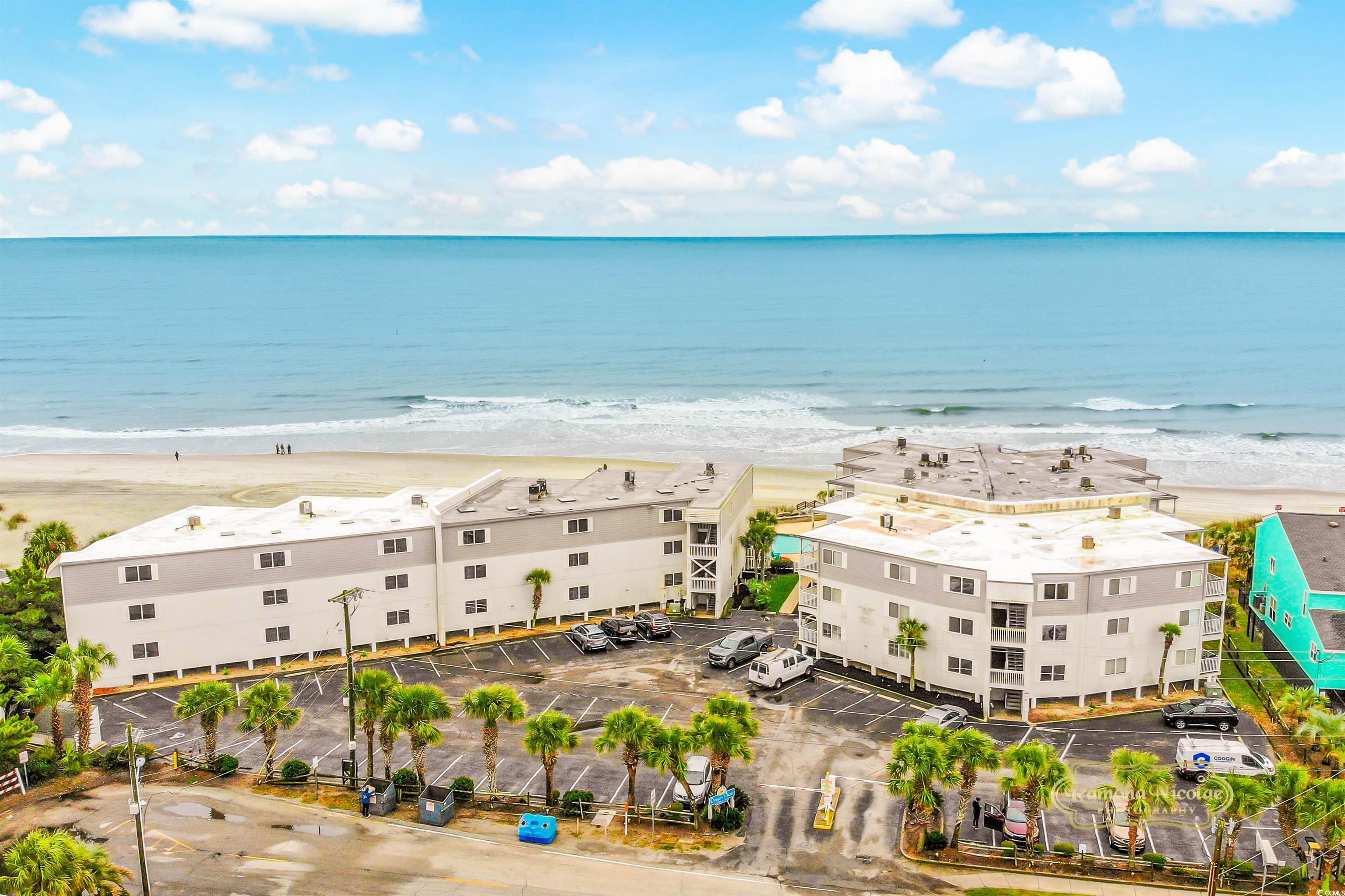 Beachfront condominium buildings and palm-lined parking lot beside a wide sandy beach and calm ocean.