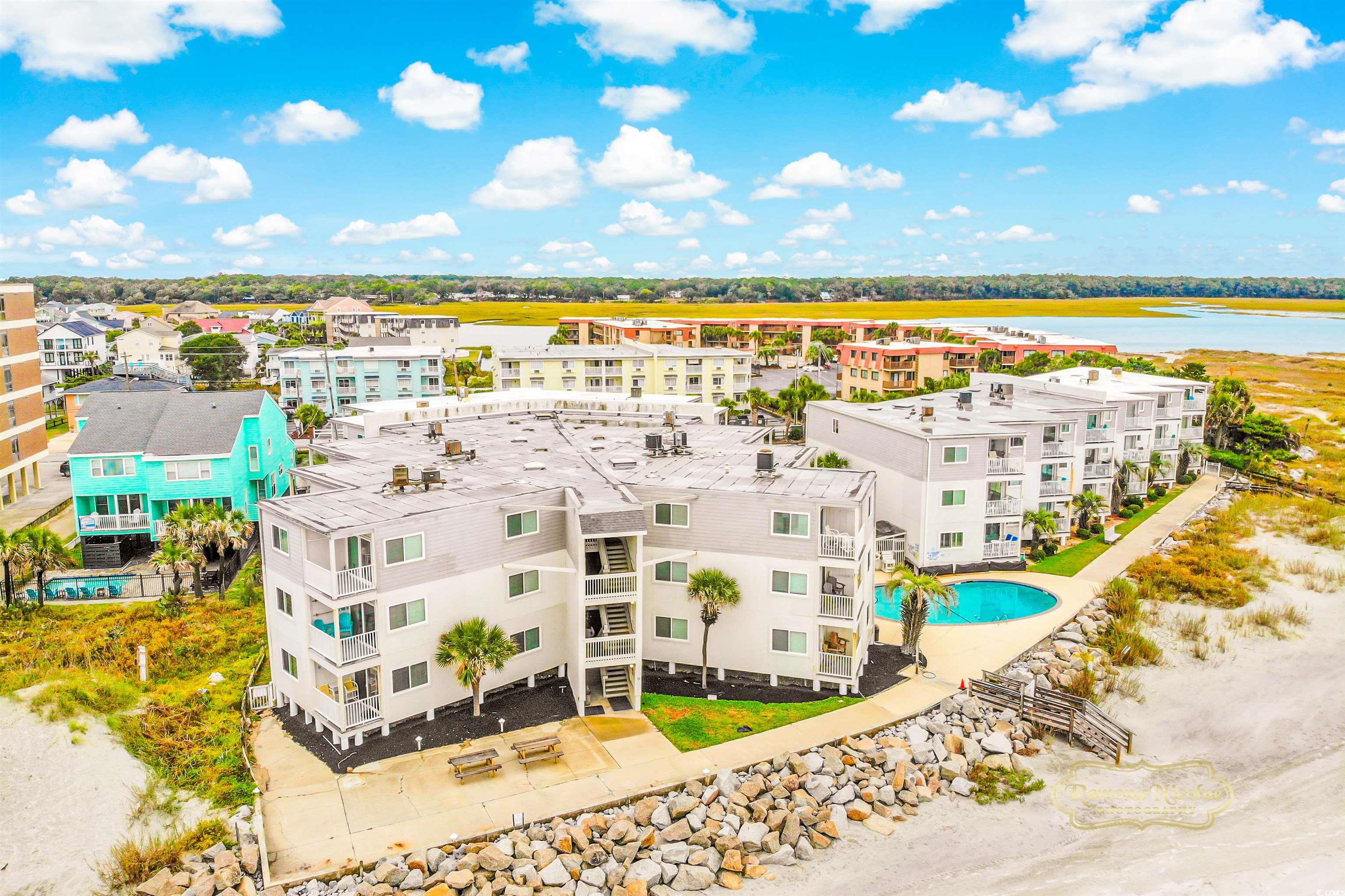 Aerial view of coastal condominiums beside a pool, sandy shoreline, and marsh water.
