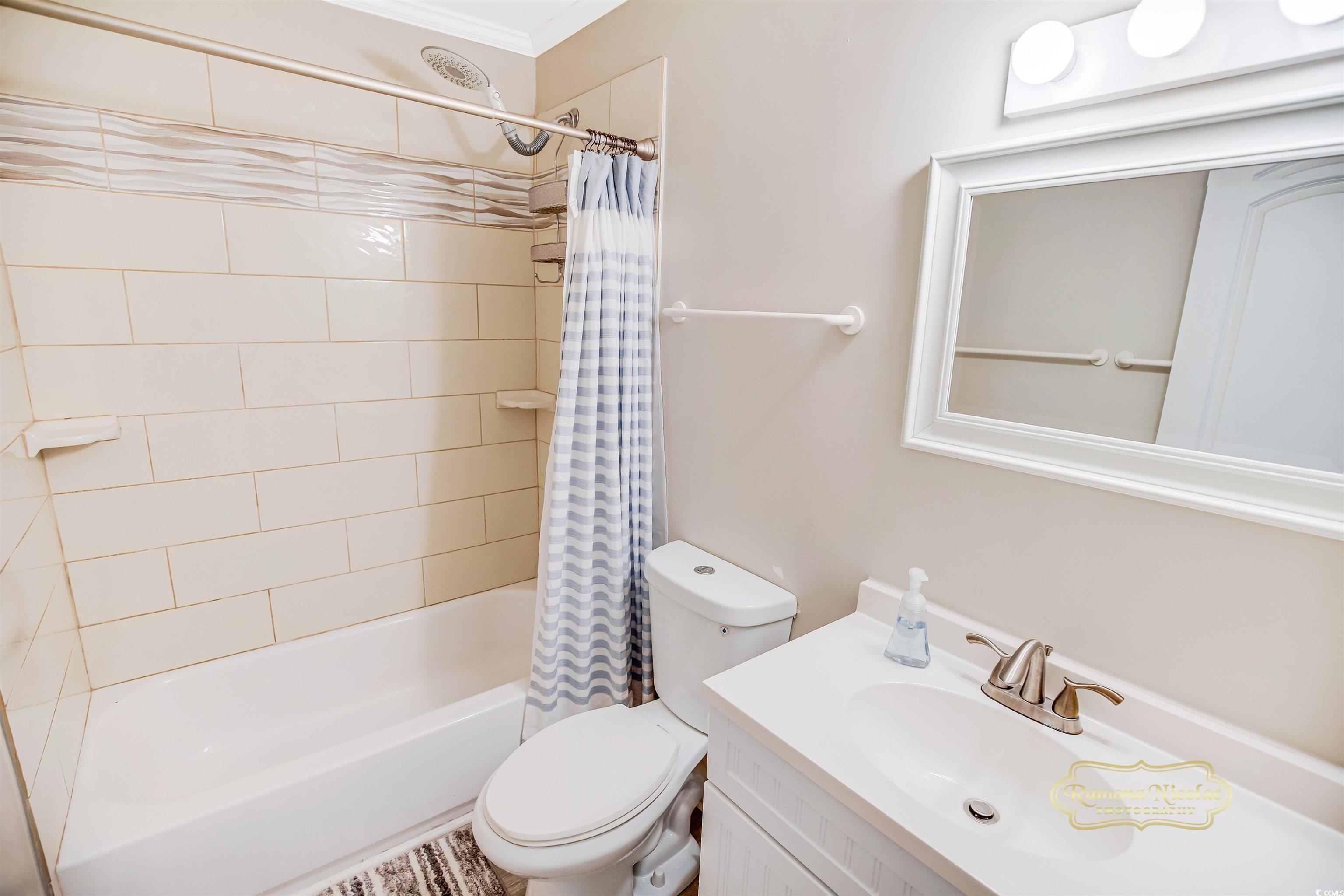 Bathroom with a tiled tub and shower, striped curtain, toilet, and white vanity.