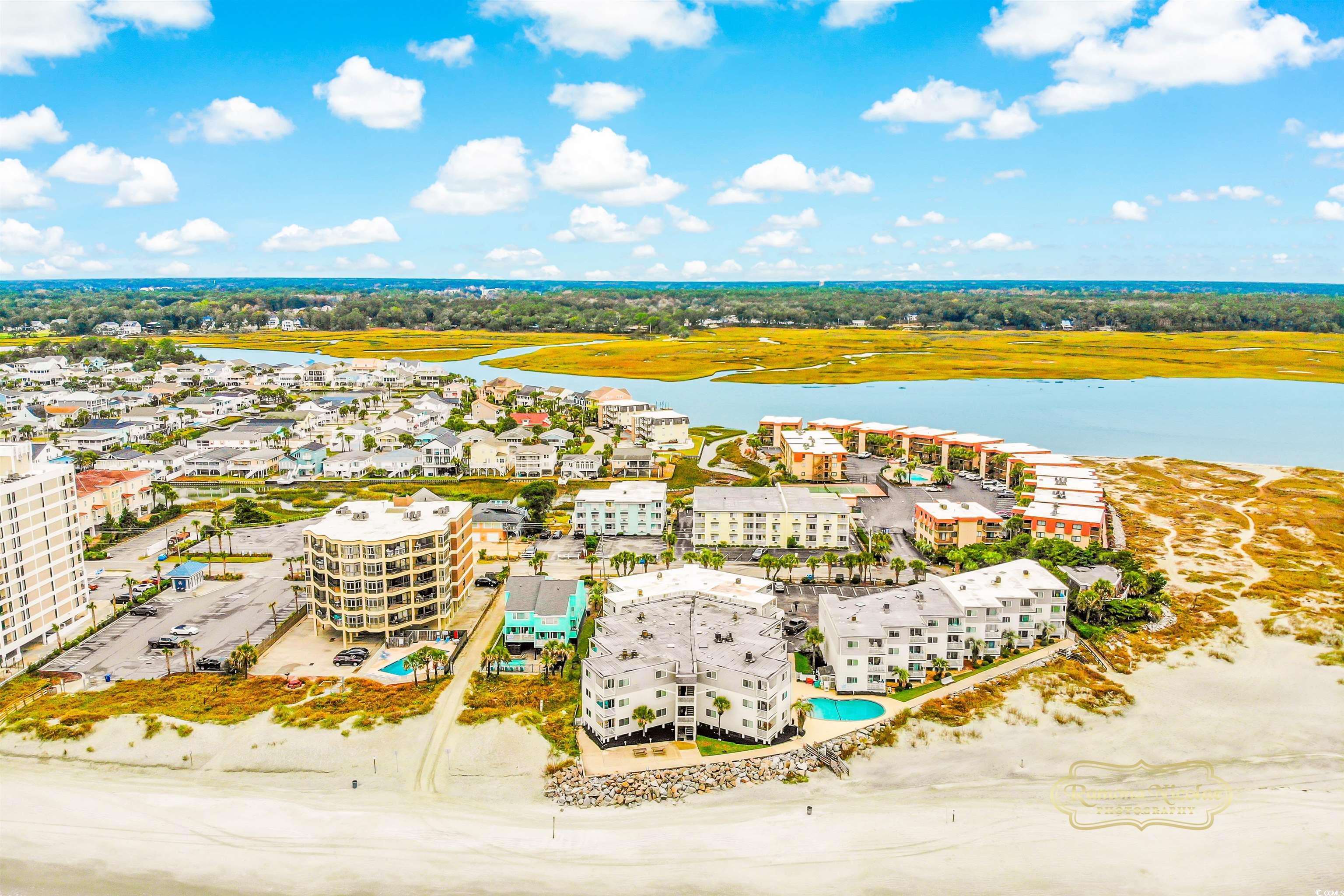 Aerial view of beachfront condos beside a sandy shore, coastal inlet, and golden marshland.