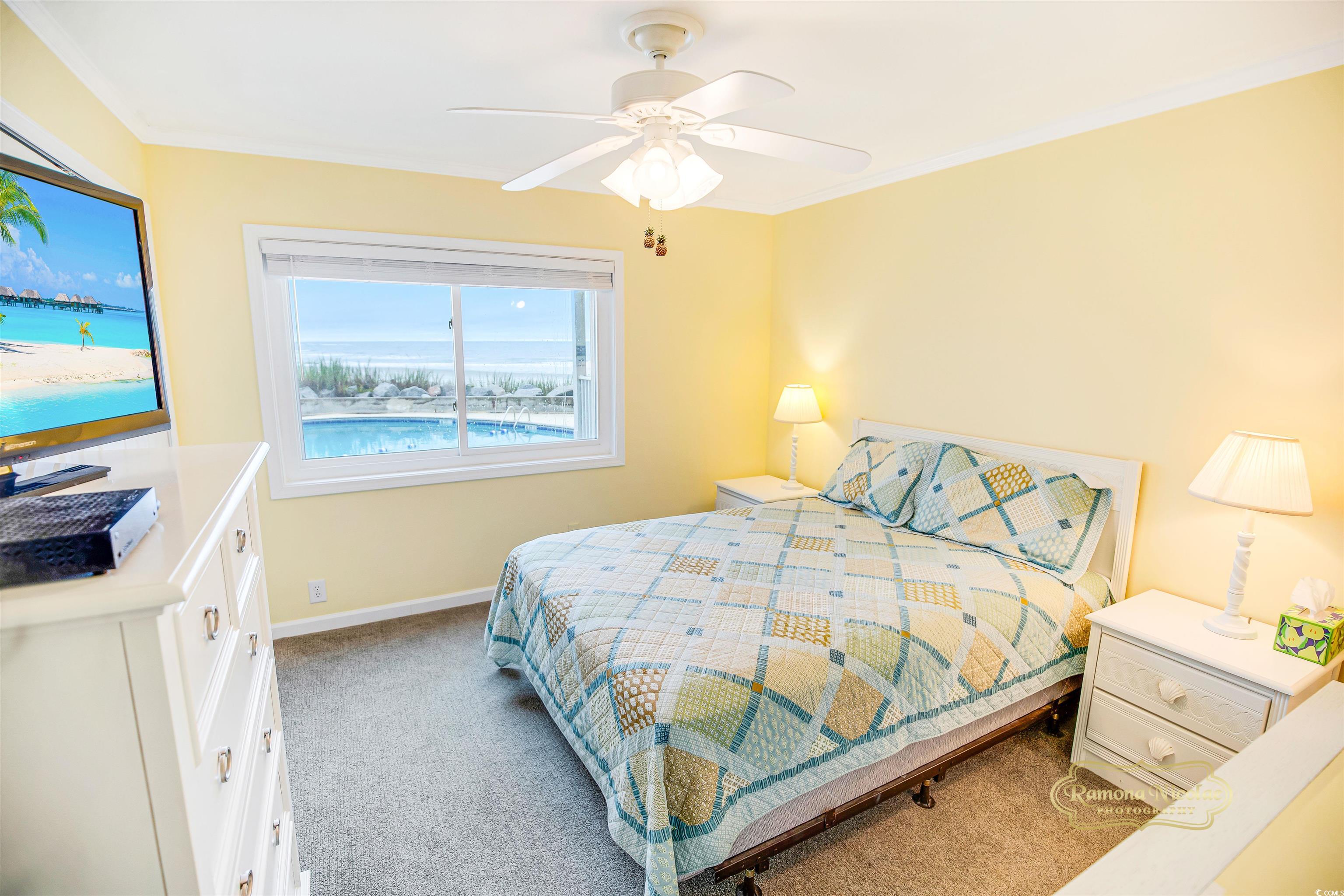 Furnished bedroom with a patchwork bedspread and a window overlooking a pool, beach, and ocean.