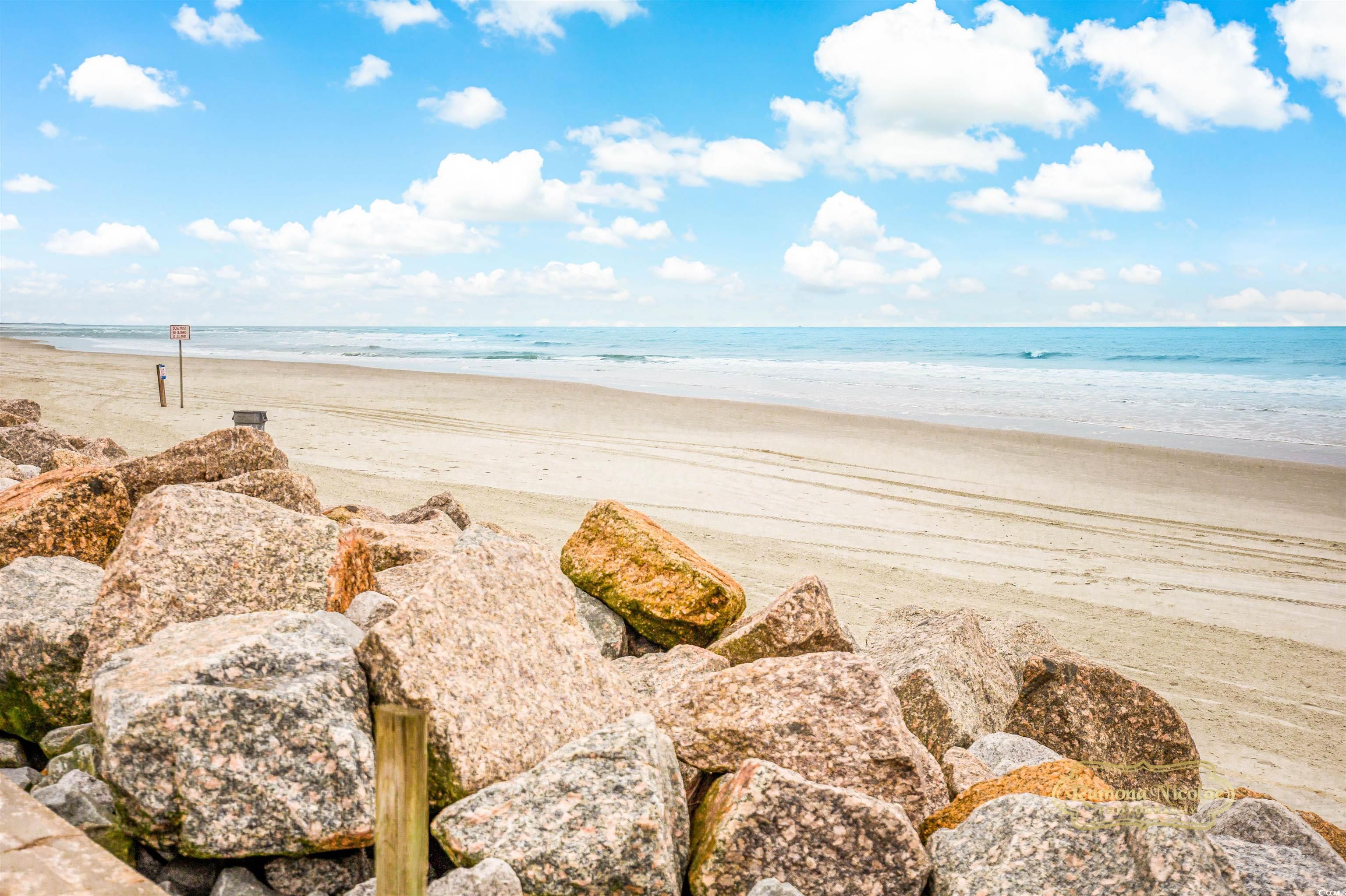 Sandy beach and calm ocean beyond foreground rocks under a bright, cloud-filled sky.