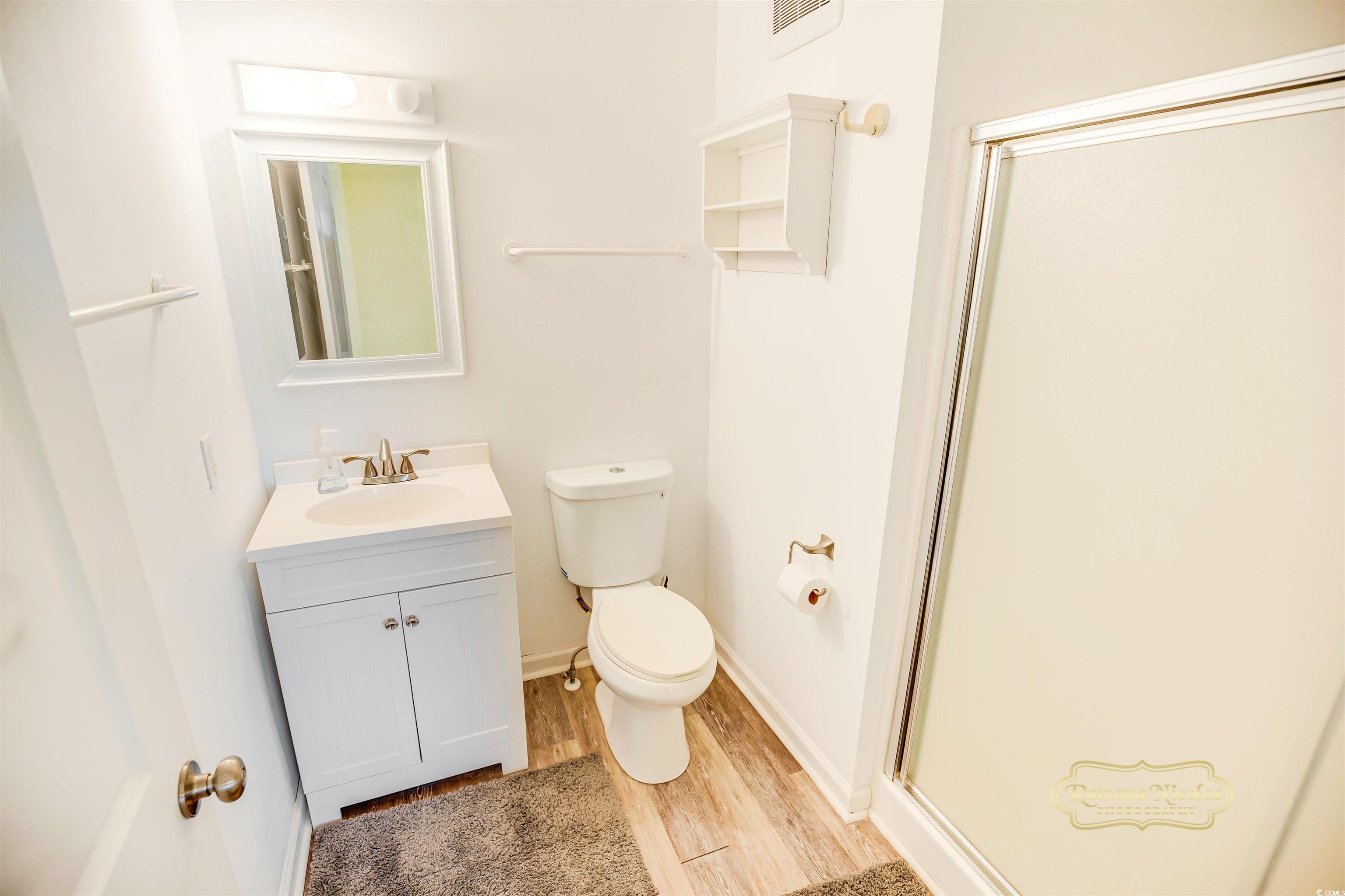 Bathroom with a white vanity, toilet, glass-enclosed shower, and wood-look flooring.