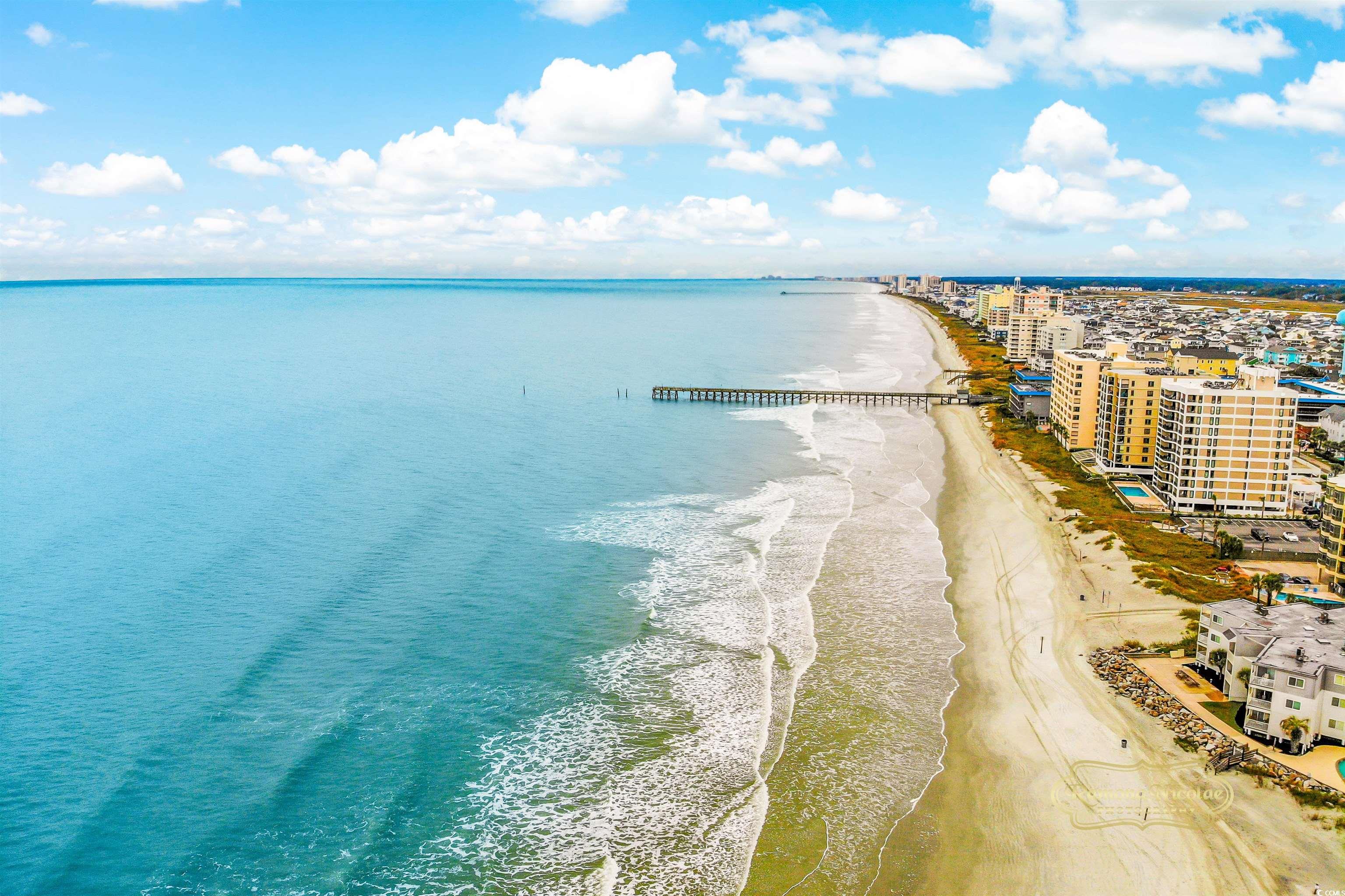 Aerial view of beachfront condominiums beside a broad sandy beach, turquoise ocean, and long pier.