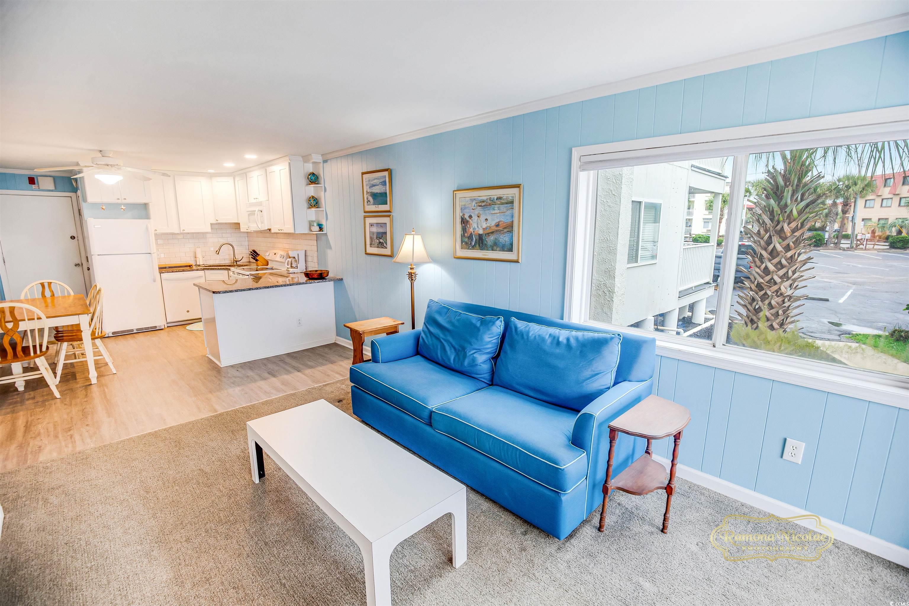 Condo living room with a bright blue sofa, open kitchen, and large window overlooking a palm tree.
