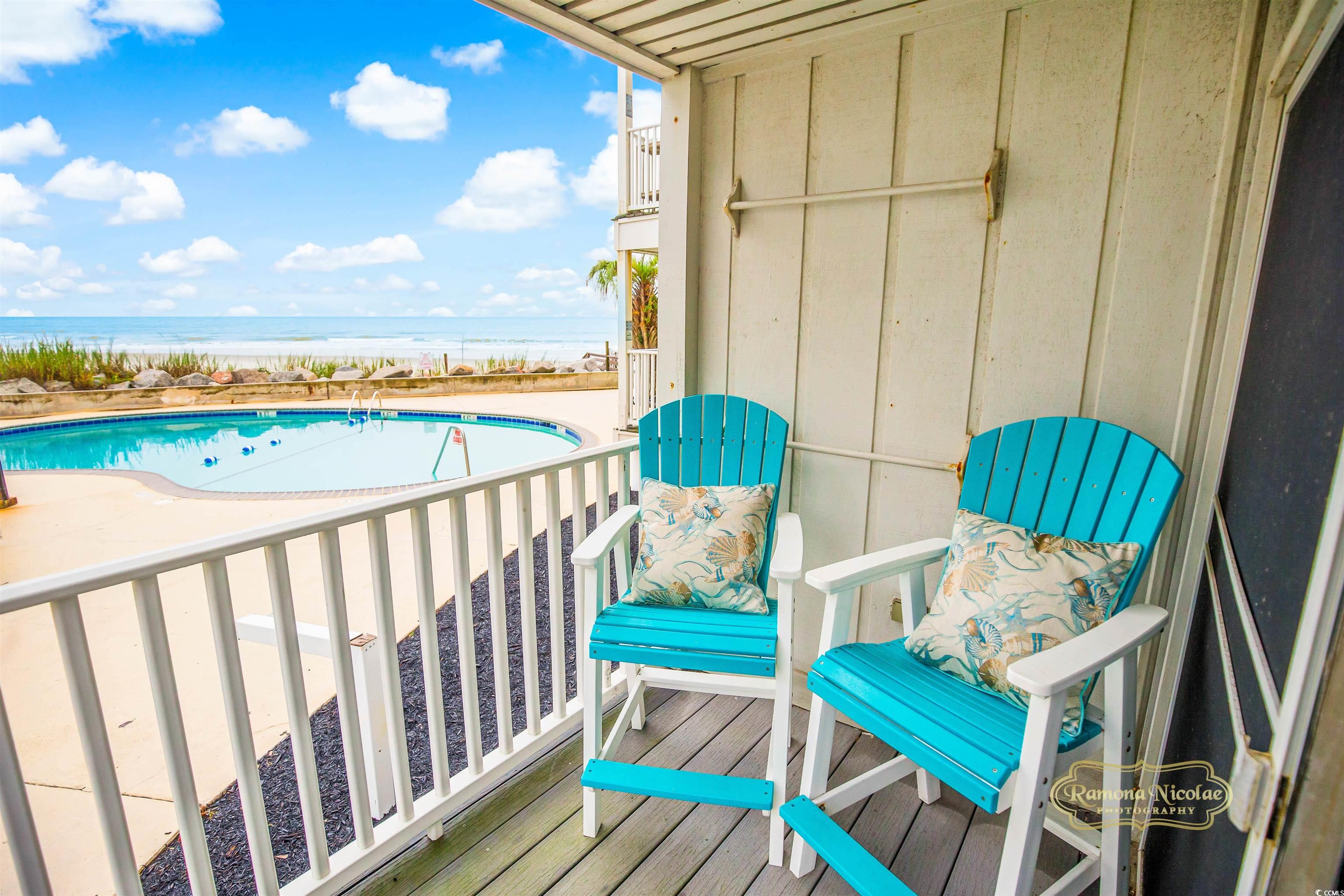 Beachfront balcony with turquoise chairs overlooking a pool, sandy beach, and ocean.