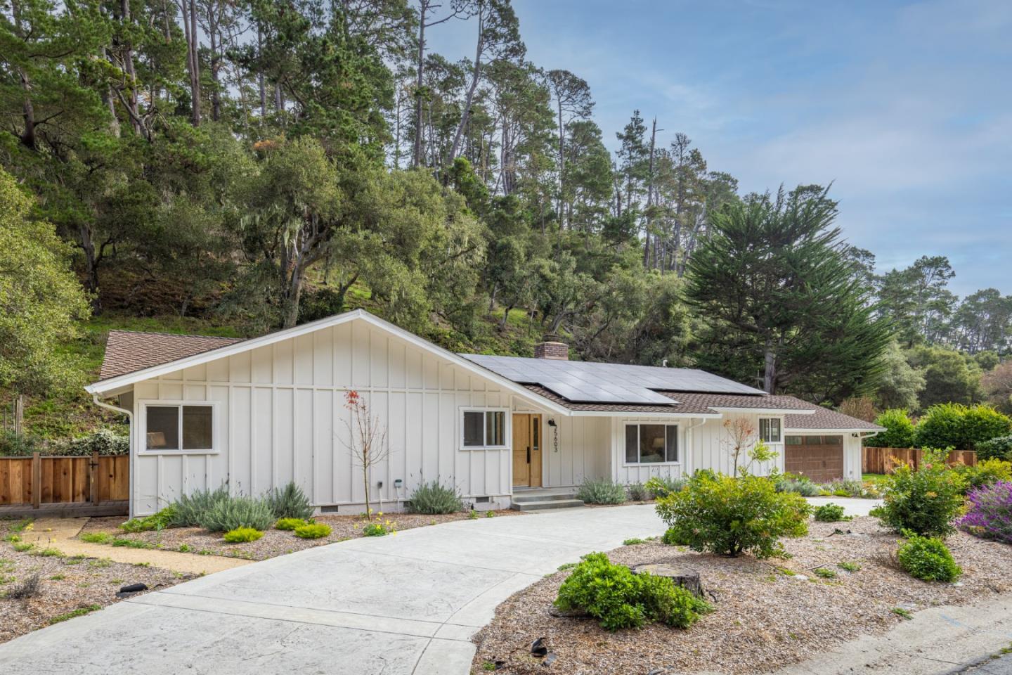 White single-story house with rooftop solar panels, a wide driveway, and a wooded hillside behind it.