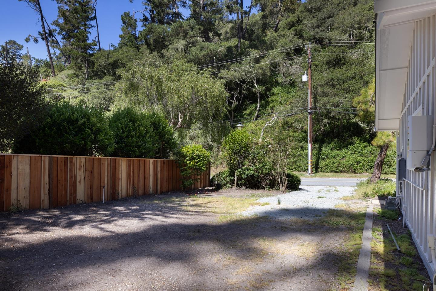 Gravel side yard beside a house, bordered by a wooden fence and dense trees.