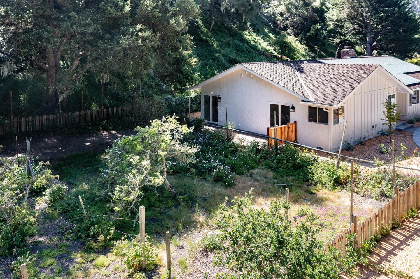 White single-story house beside a fenced, overgrown garden and wooded hillside.