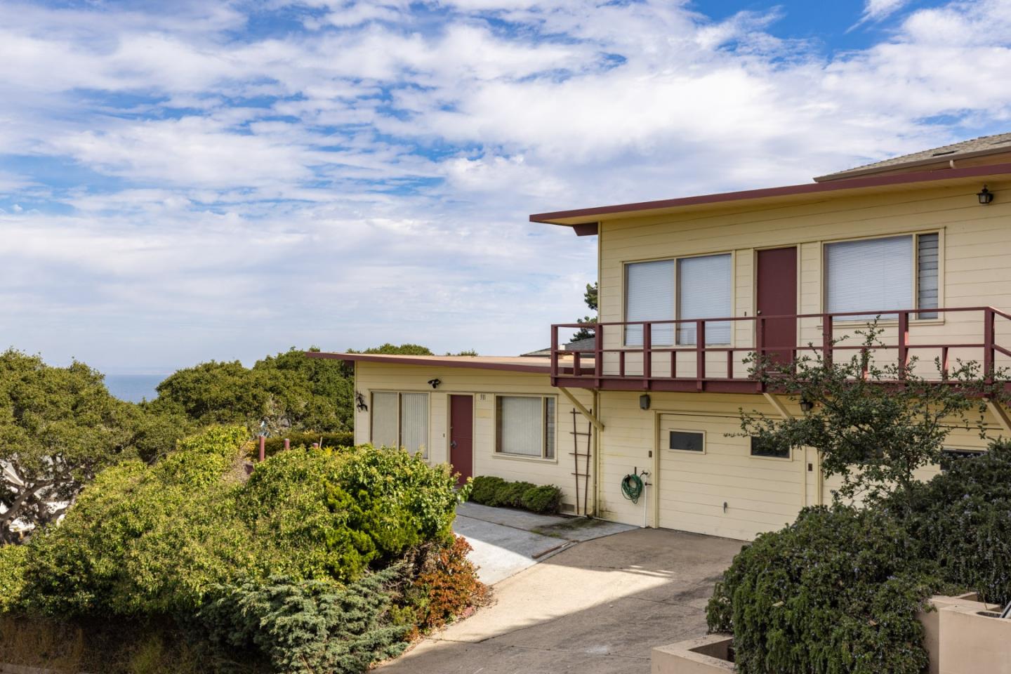 Pale yellow two-story house with a burgundy balcony, driveway, and ocean glimpsed through trees.
