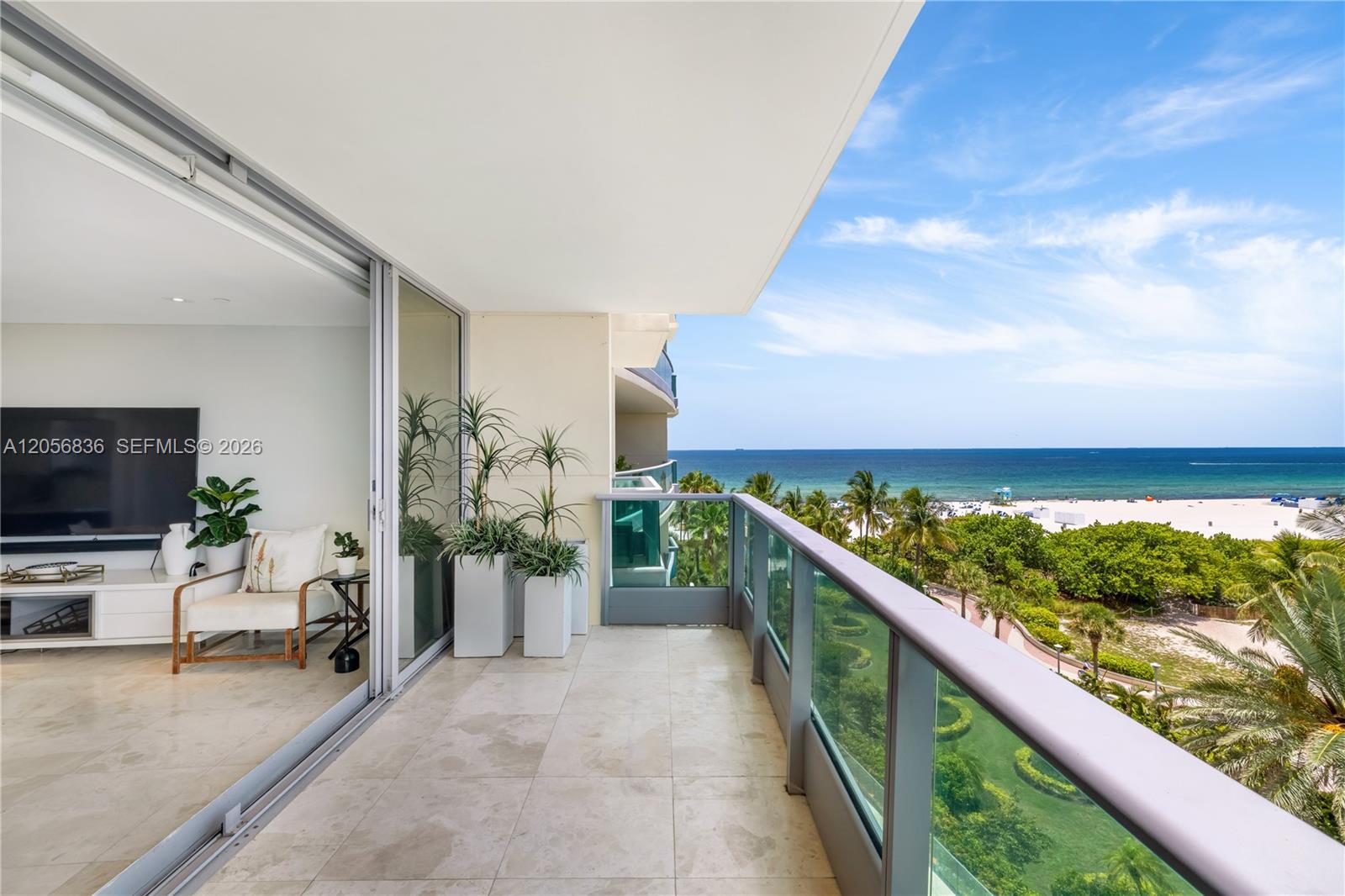 Condominium balcony overlooking the beach and ocean, with glass railings and a furnished living room visible inside.