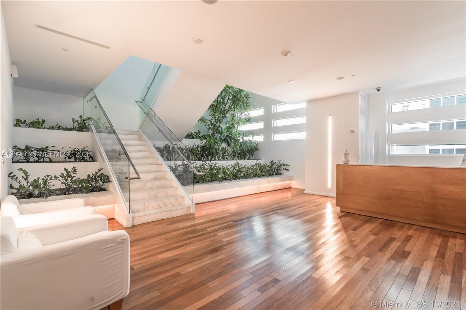 Condominium lobby with a glass-railed staircase, indoor greenery, and a wood reception desk.