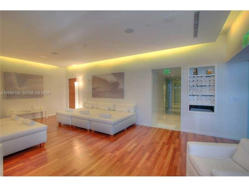 Furnished lounge with white sectional sofas, warm wood floors, and recessed ceiling lighting.